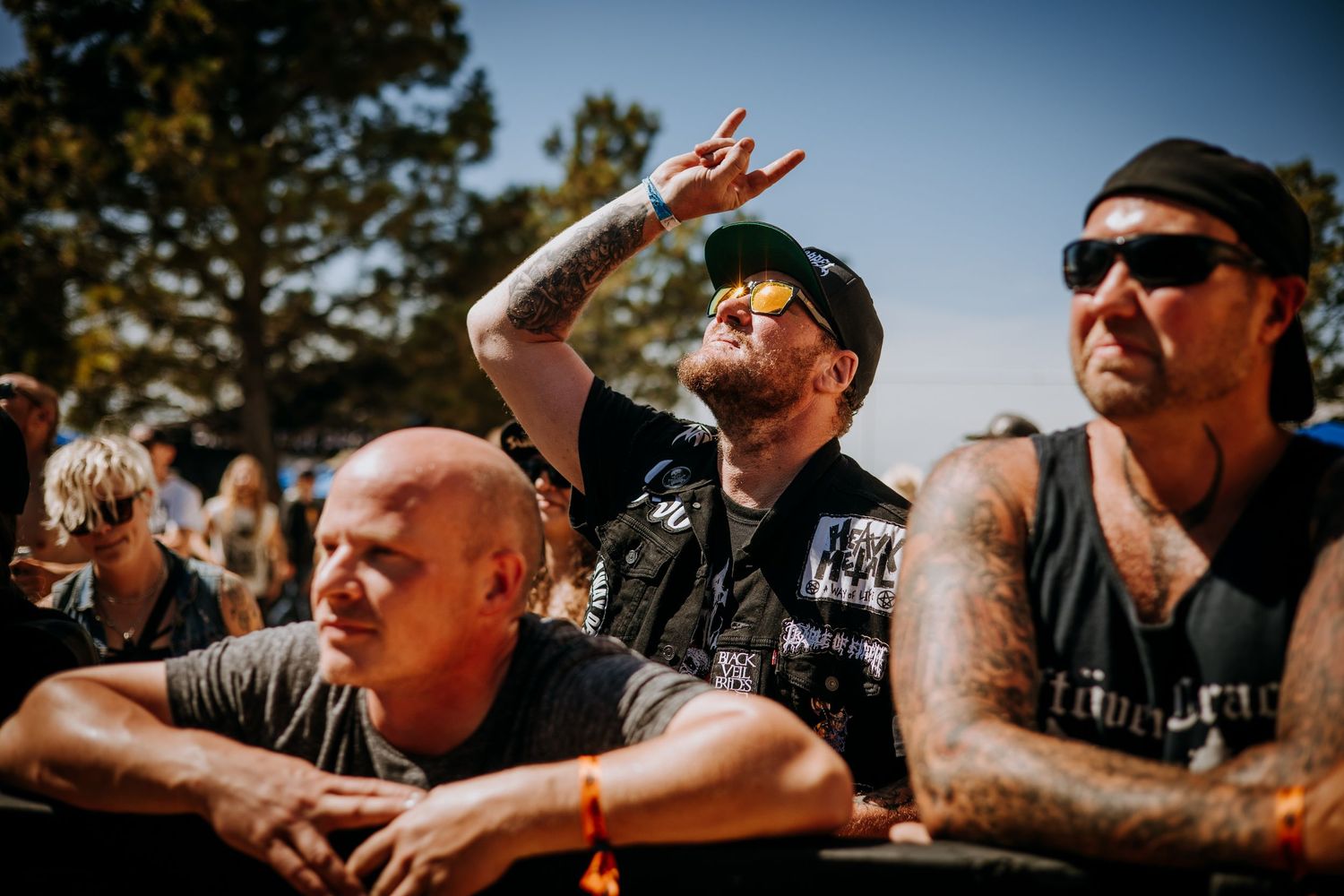 Energetic crowd members with tattoos and sunglasses cheering enthusiastically at an outdoor music festival.