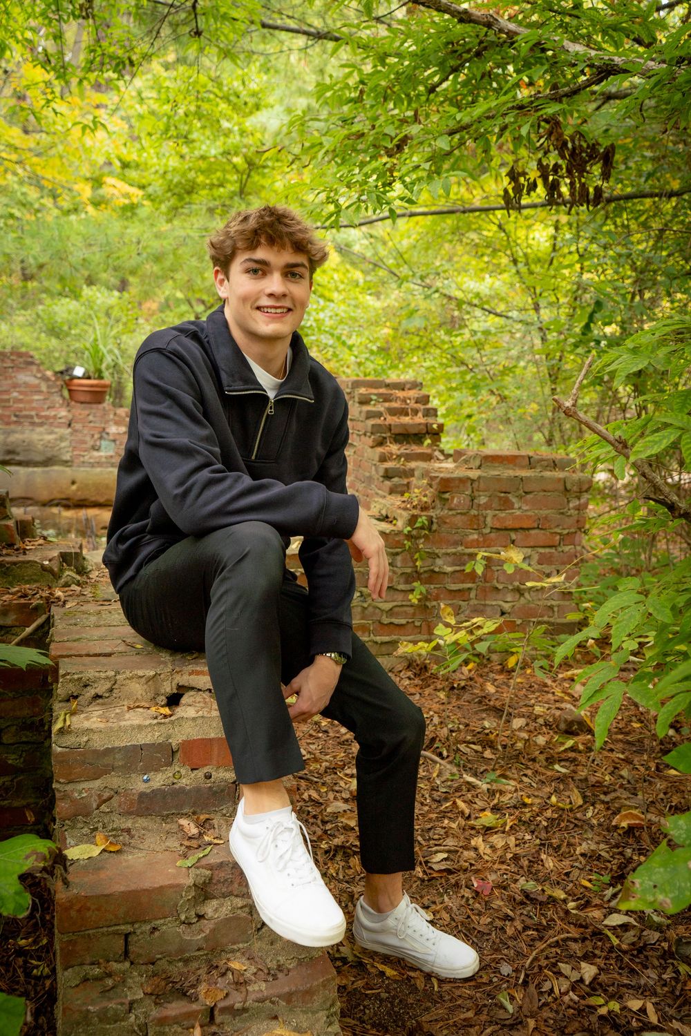 Person sitting casually on vintage brick wall surrounded by lush green foliage in outdoor setting.