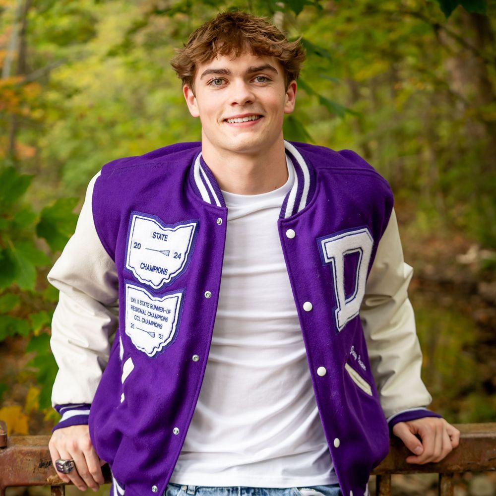 A person wearing a purple and white varsity letterman jacket sits casually outdoors against a natural backdrop.
