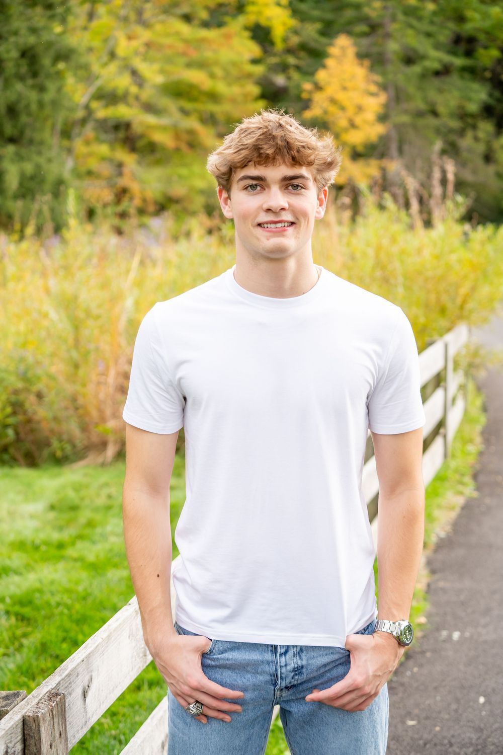 Person in a white t-shirt and blue jeans poses in a scenic outdoor setting with autumn foliage in the background.