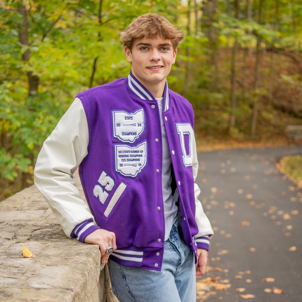 Student sitting on stone wall wearing purple varsity jacket surrounded by autumn foliage.
