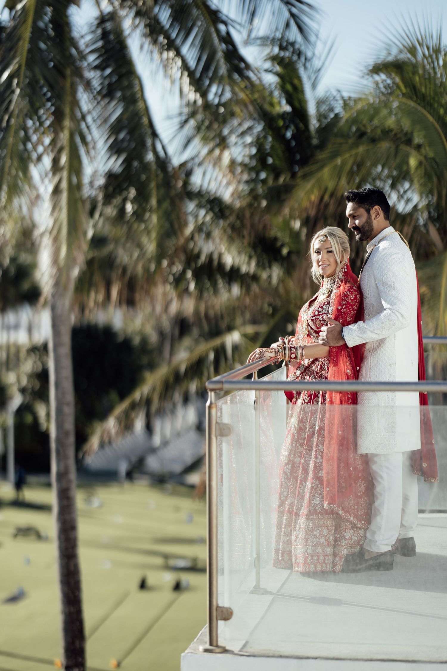 A couple in traditional wedding attire embraces on a balcony surrounded by palm trees in a park setting.