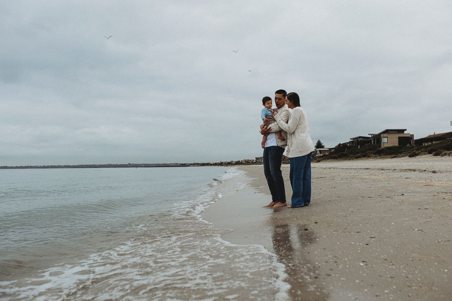 Family captured during a family photography session playing along a beach shoreline under a cloudy sky on a calm day.