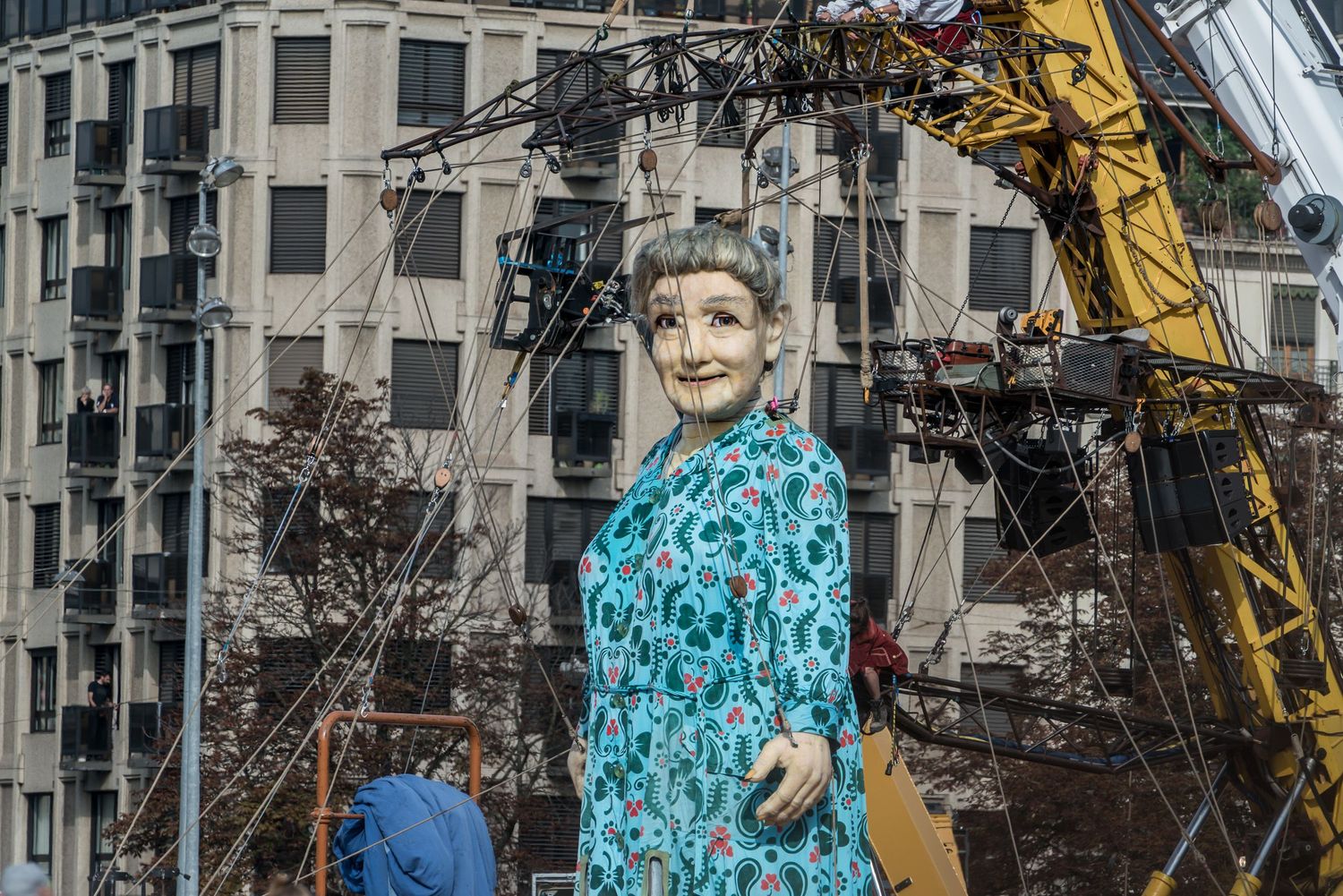 A towering figure in a turquoise patterned dress stands against an urban backdrop with construction equipment nearby.