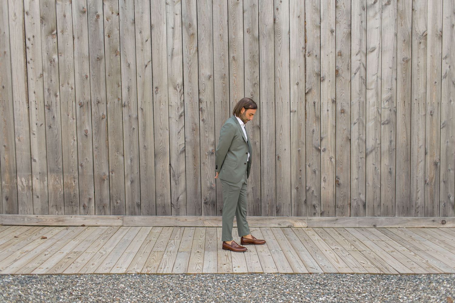 A person in a gray suit stands on a wooden deck against a weathered wooden fence.