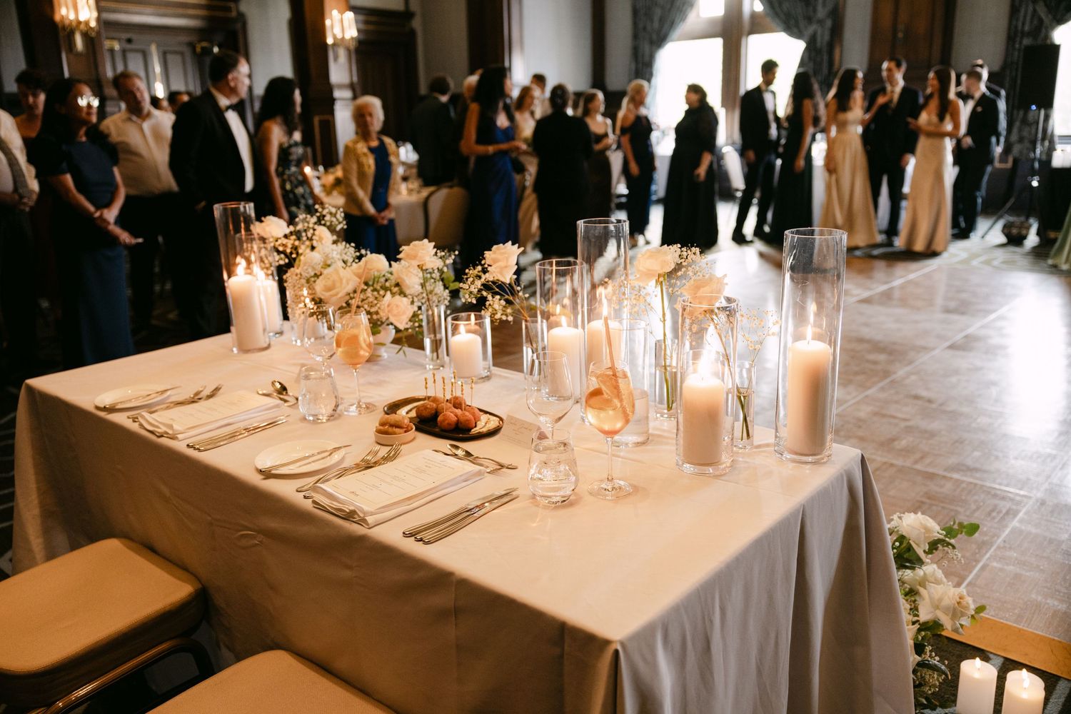 Elegant wedding reception table setting with glowing candles and formal place settings in an ornate venue with guests mingling.