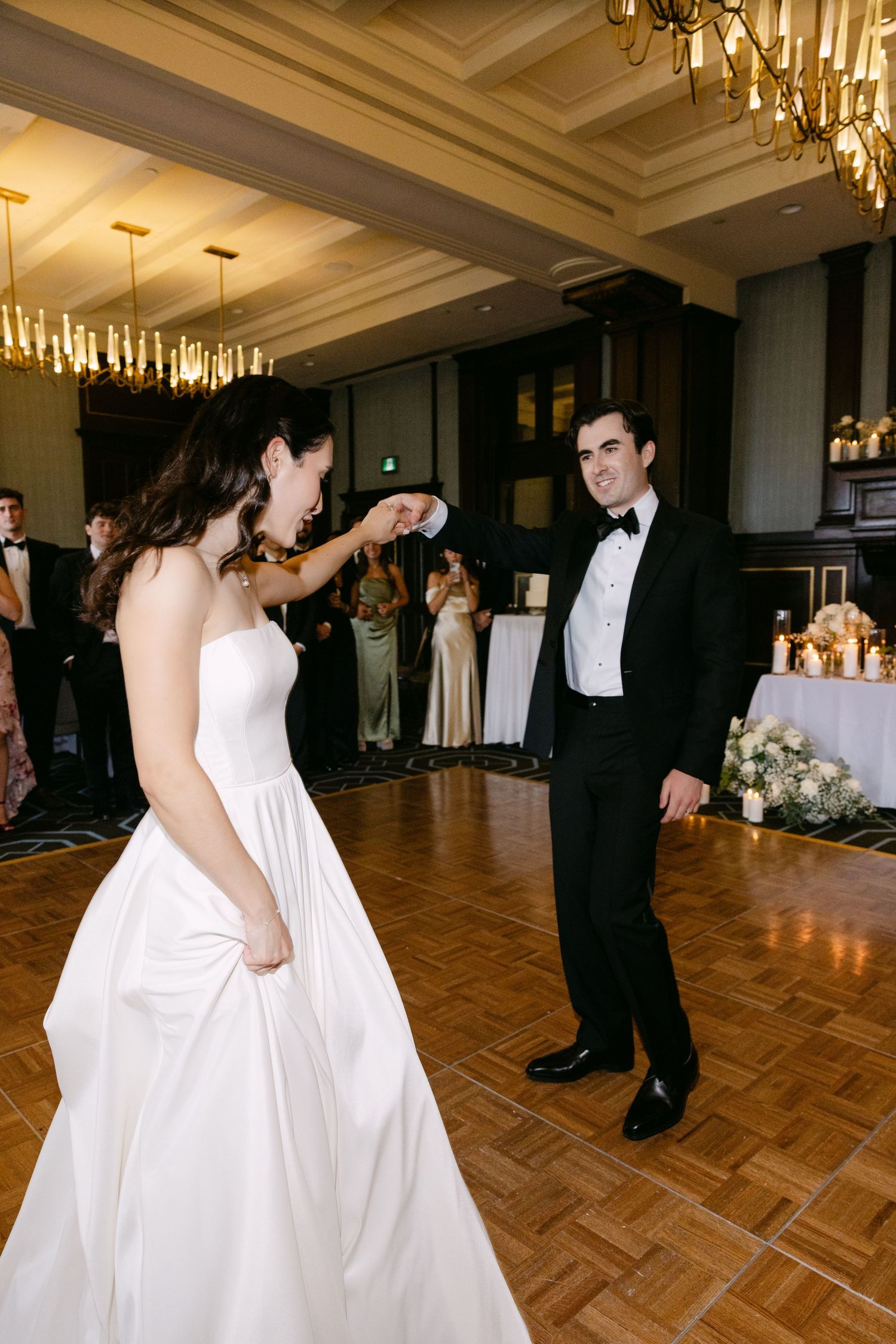 A couple shares their first dance on a parquet floor at an elegant wedding reception with crystal chandeliers overhead.