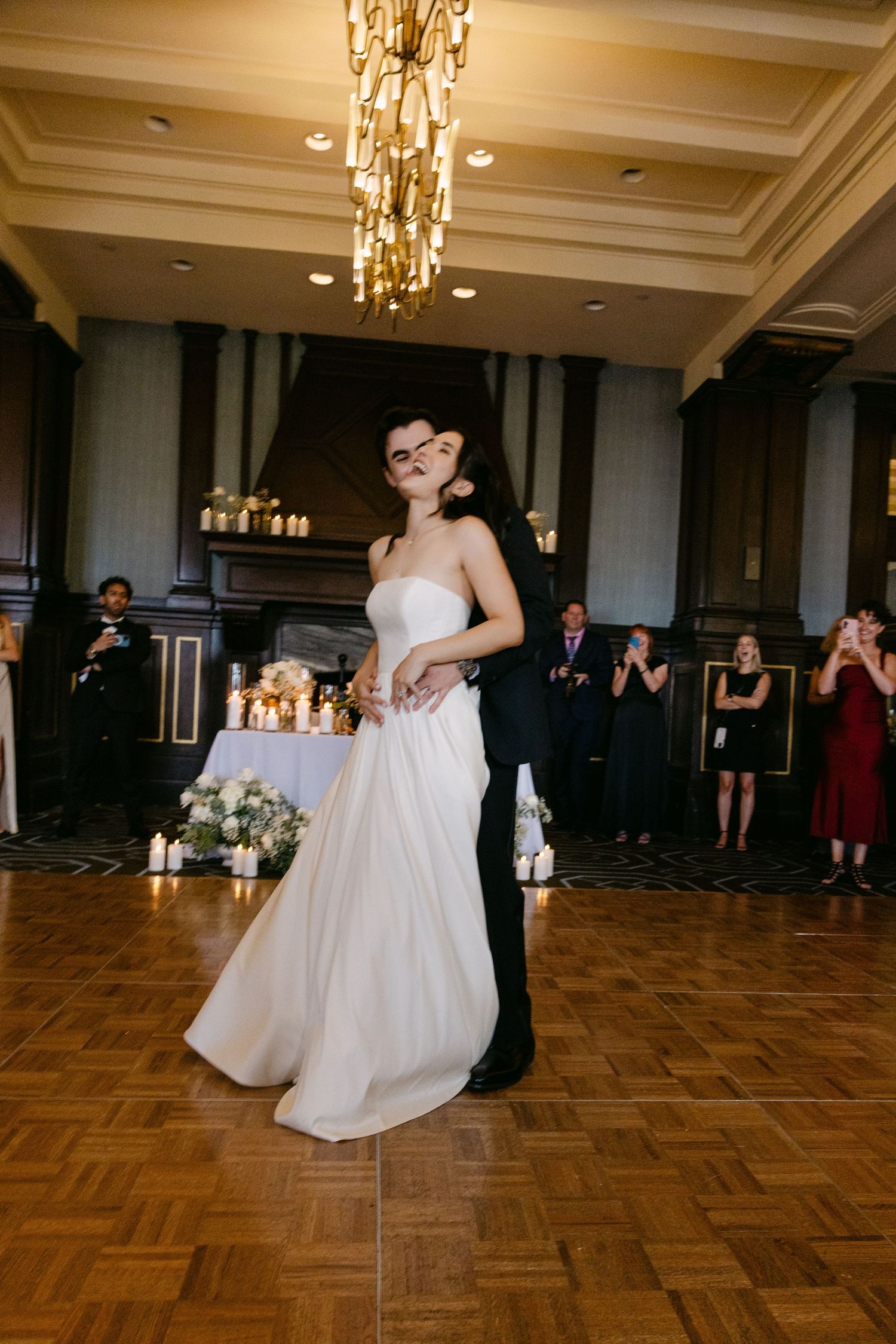 Couple sharing their first dance at an elegant wedding reception with parquet flooring and crystal chandeliers.