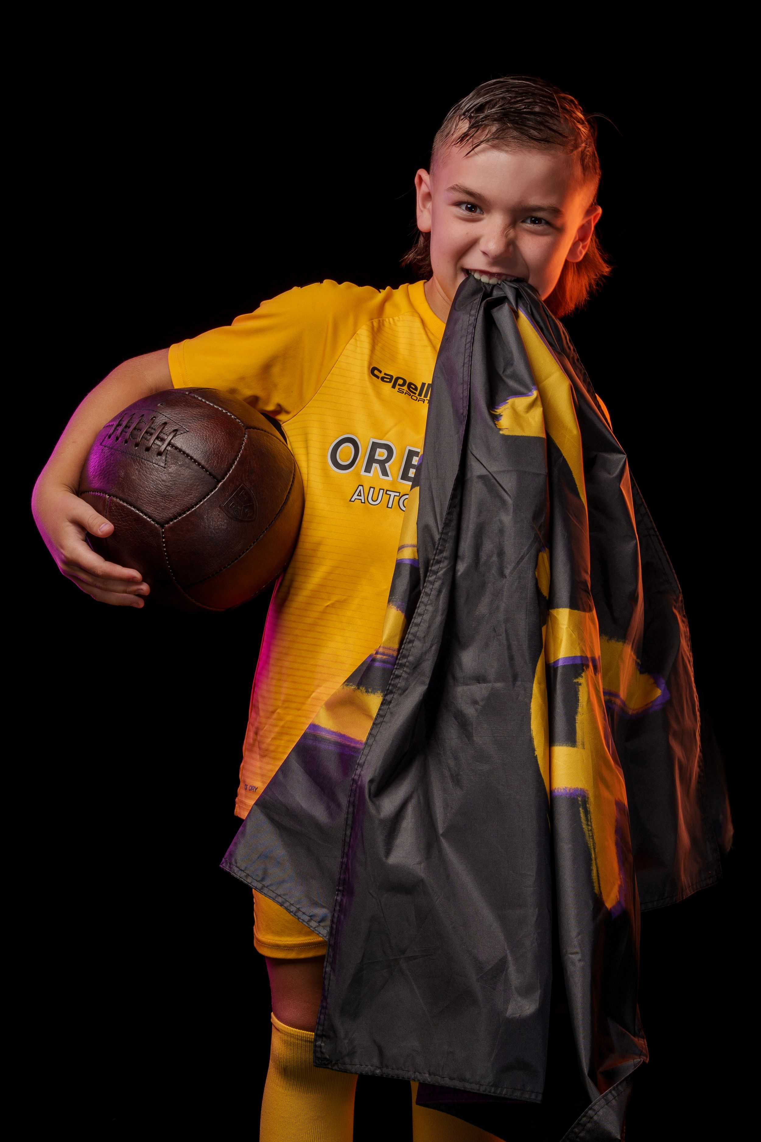 Soccer player in yellow jersey holds football and grey cape in dramatic studio lighting.