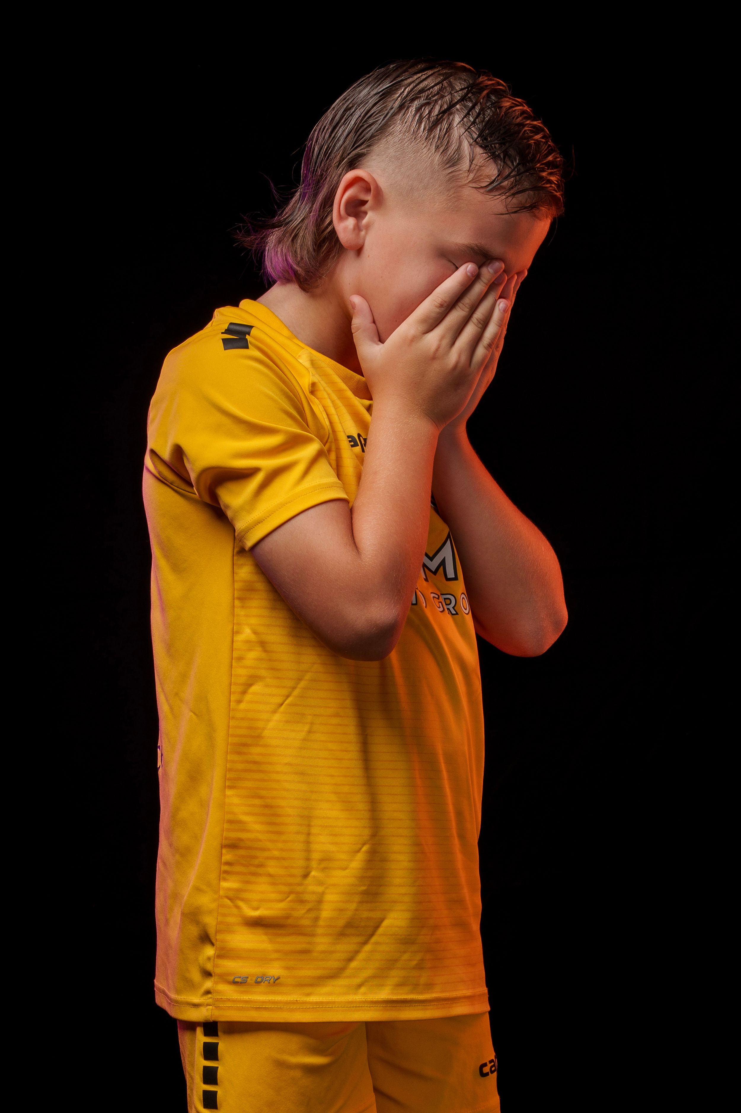 Young athlete in yellow uniform covers face with hands against dark background.
