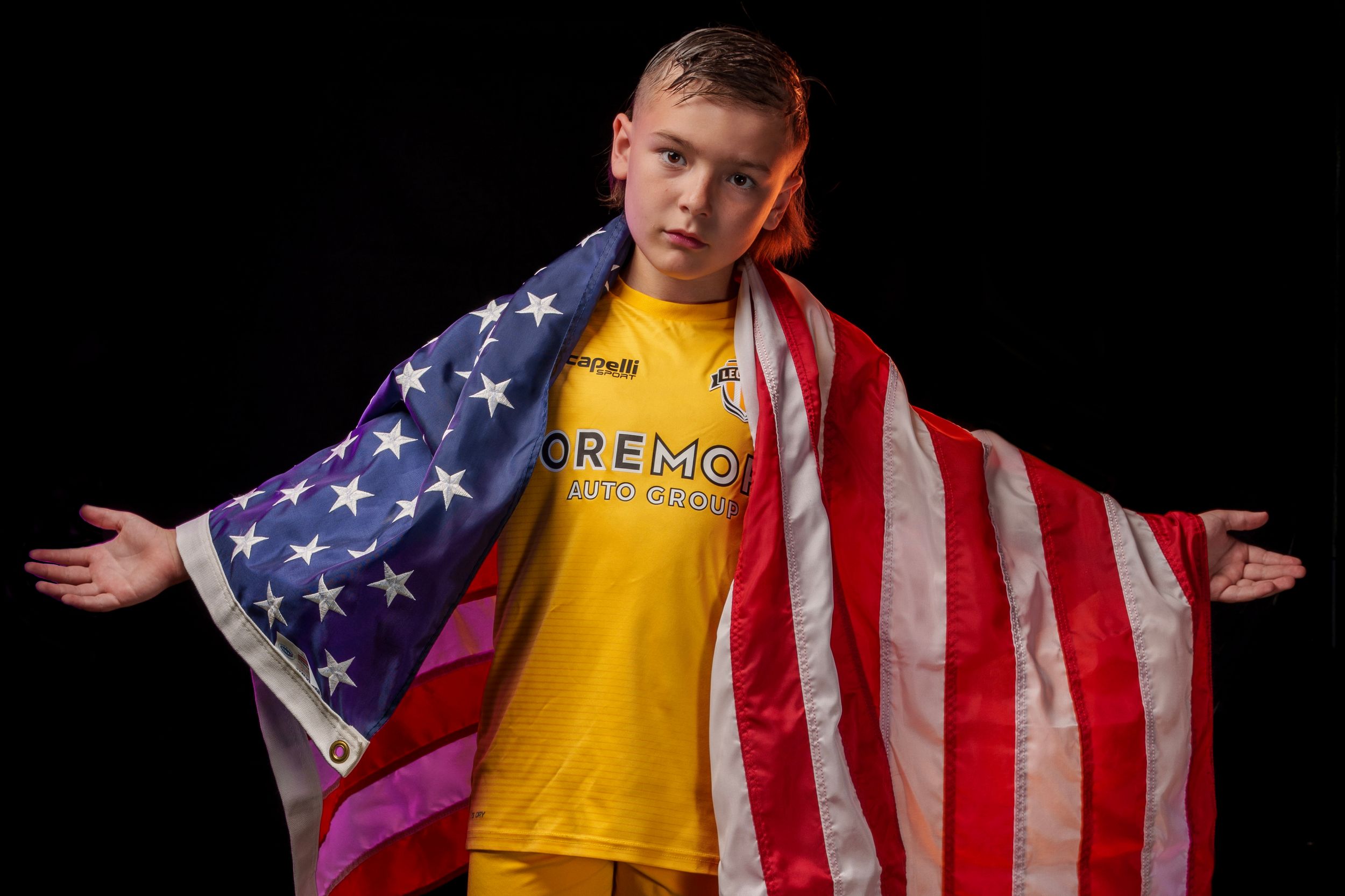Athlete in yellow Legends FC uniform poses dramatically with American flag as cape.