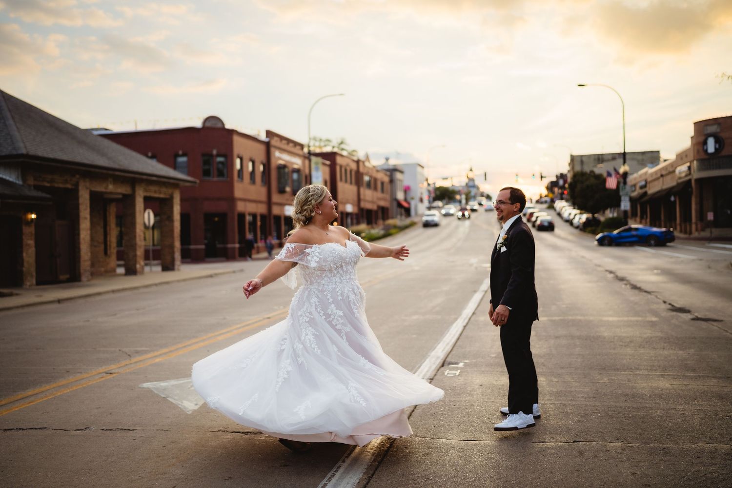 A romantic outdoor wedding photoshoot with bride in flowing white dress twirling in front of brick buildings at sunset.