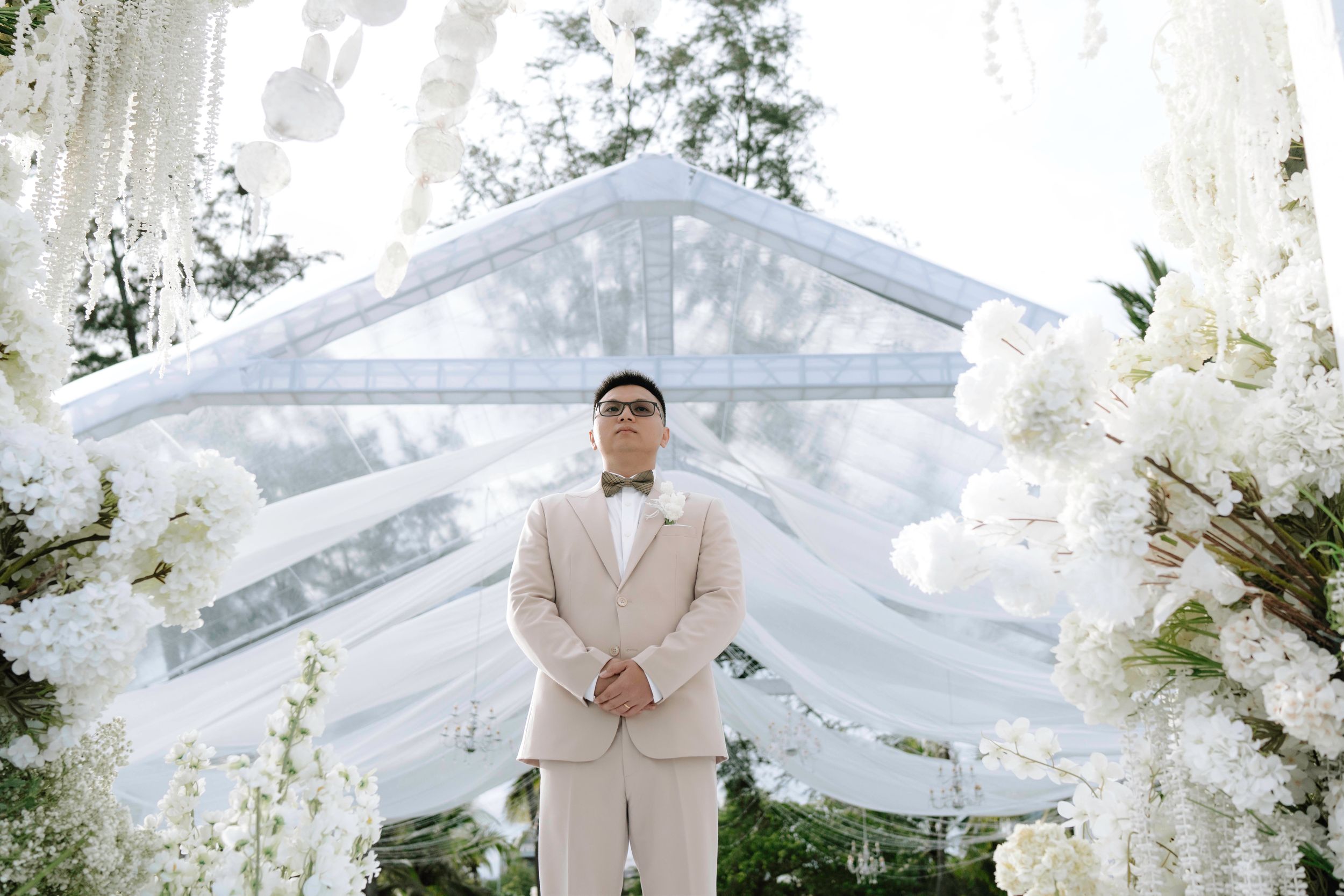 Groom in cream suit stands at glass-roofed outdoor wedding altar surrounded by white florals.