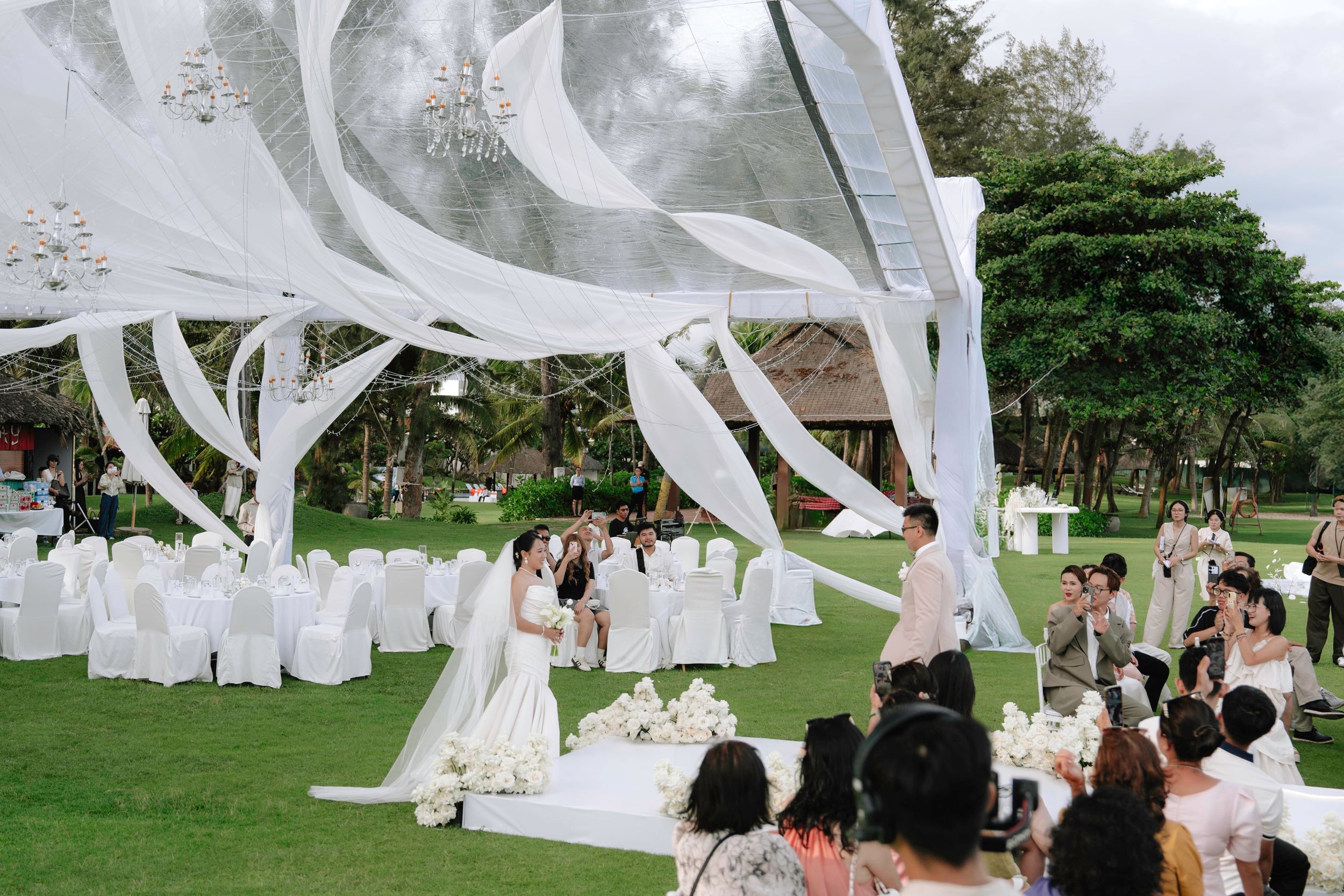 Outdoor wedding ceremony on green lawn with white draping canopy and guests seated in chairs.