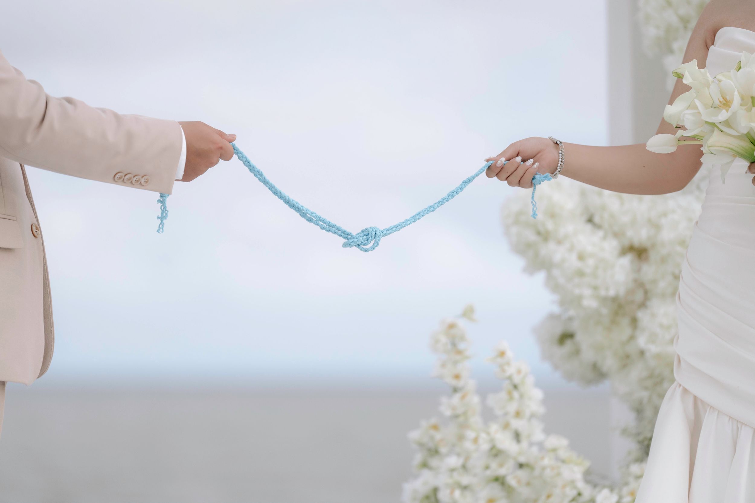 Bride and groom hold blue rope tied in knot during outdoor wedding ceremony with white florals.