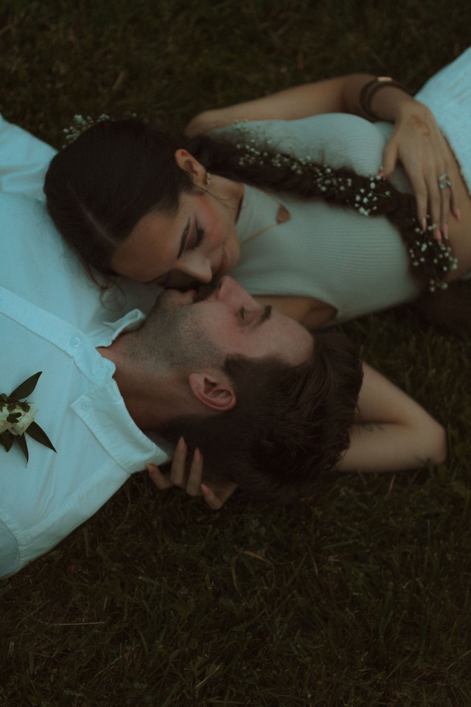 A couple in white formal attire lies peacefully on dark grass in an artistic romantic photoshoot.