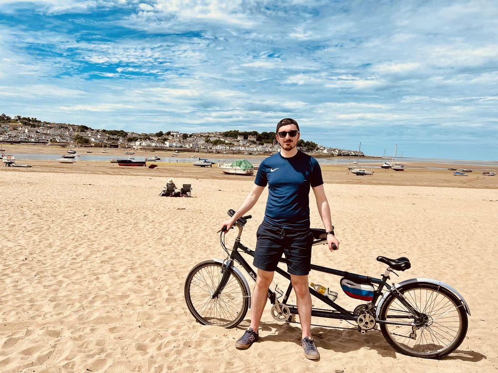 Someone poses with a silver bicycle on a sunny beach with coastal cliffs and blue skies in the background.