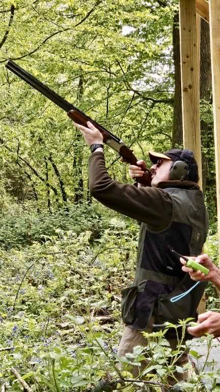 Man aiming shotgun skyward in woodland setting, wearing ear protection and shooting vest during clay pigeon shooting.