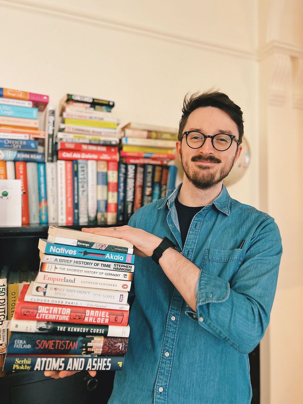 Person in denim shirt posing with stacks of books in front of bookshelf.