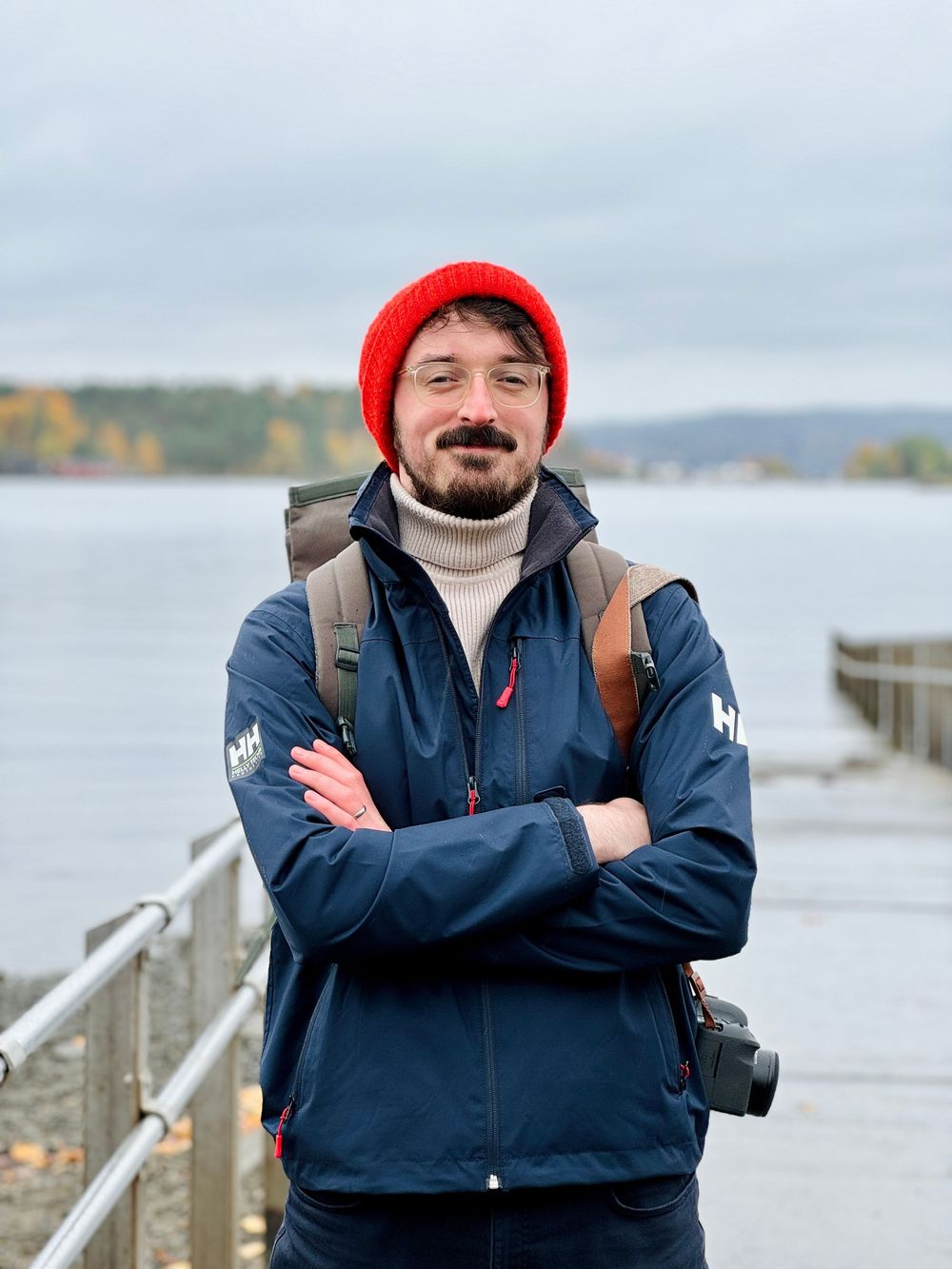 Person in navy coat and red beanie stands on wooden dock by misty waterfront.