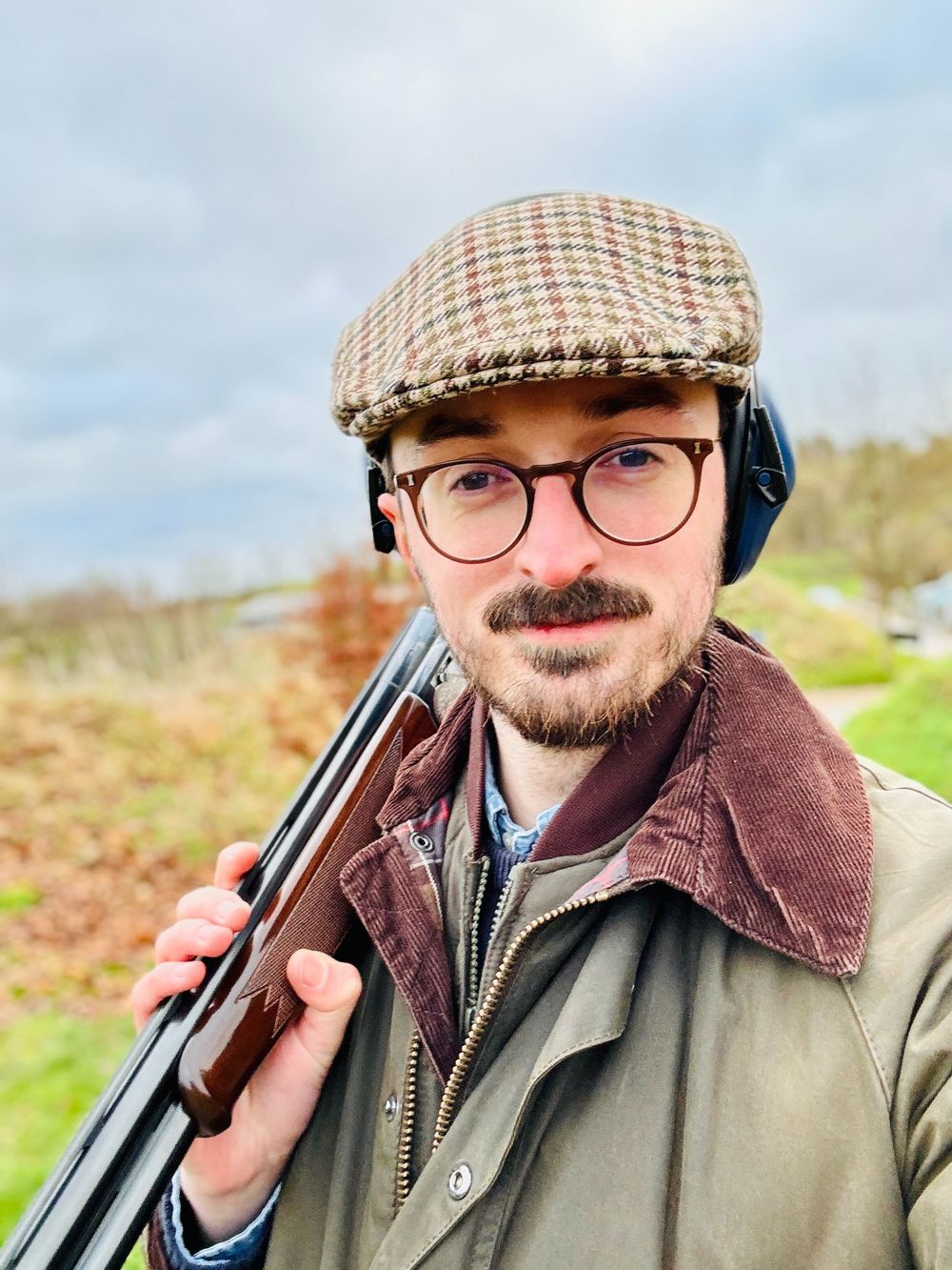 Man in tweed cap and brown jacket posing with shotgun outdoors against cloudy sky.