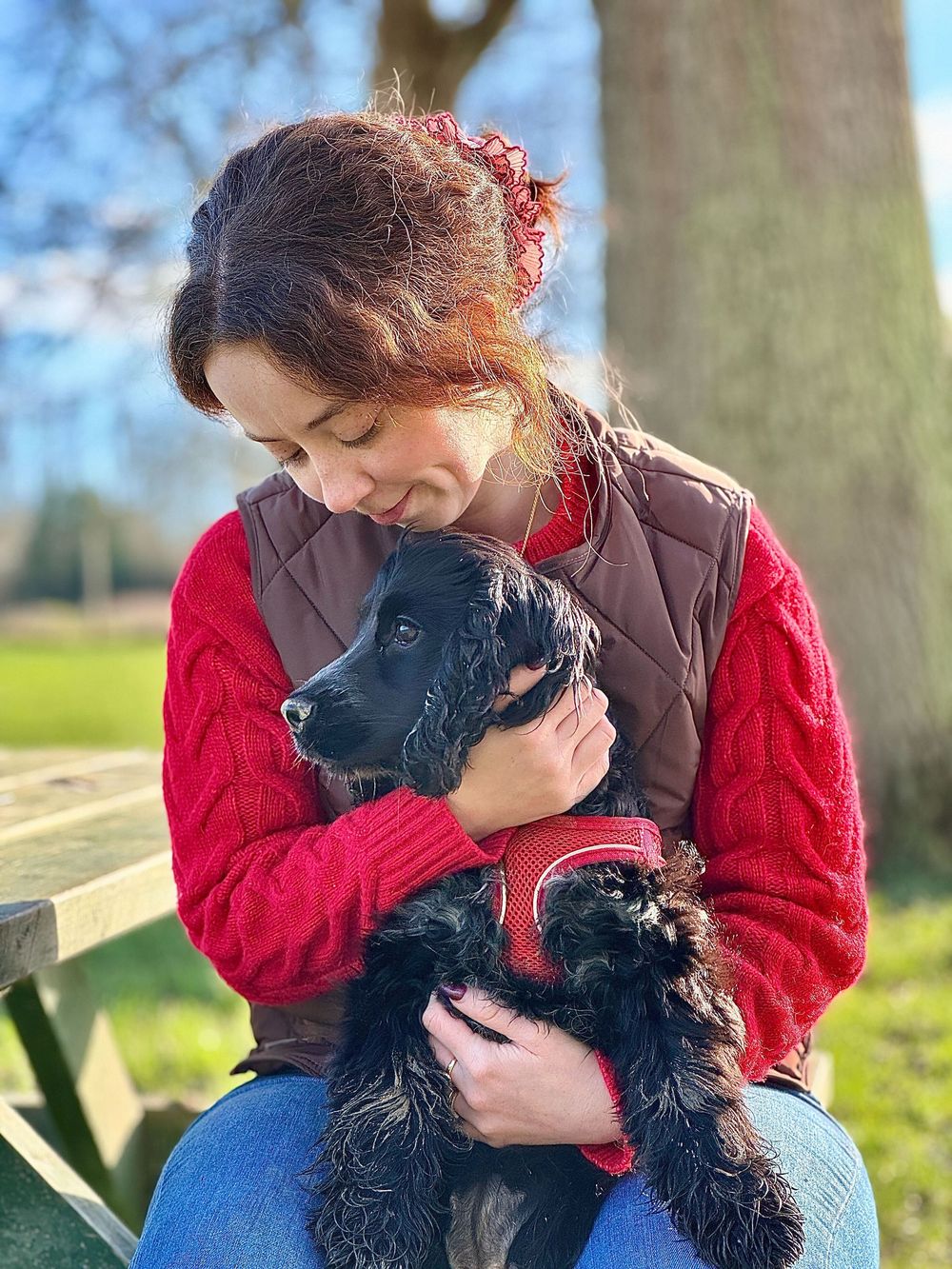 A person in a red sweater and brown jacket shares affectionate moments with a black dog in a sunny outdoor setting.