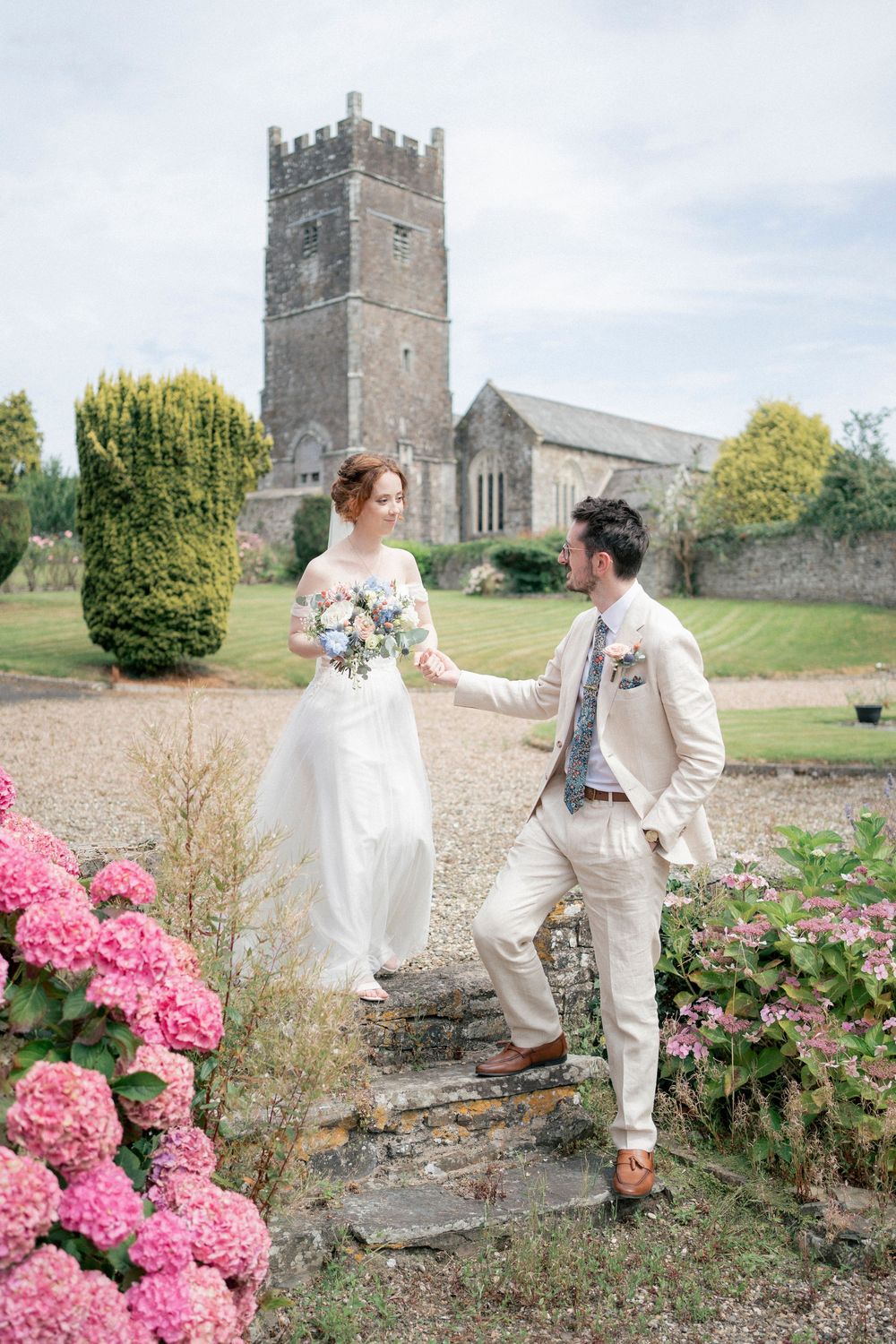 A couple poses playfully in a garden with pink hydrangeas and a historic stone church tower in the background.