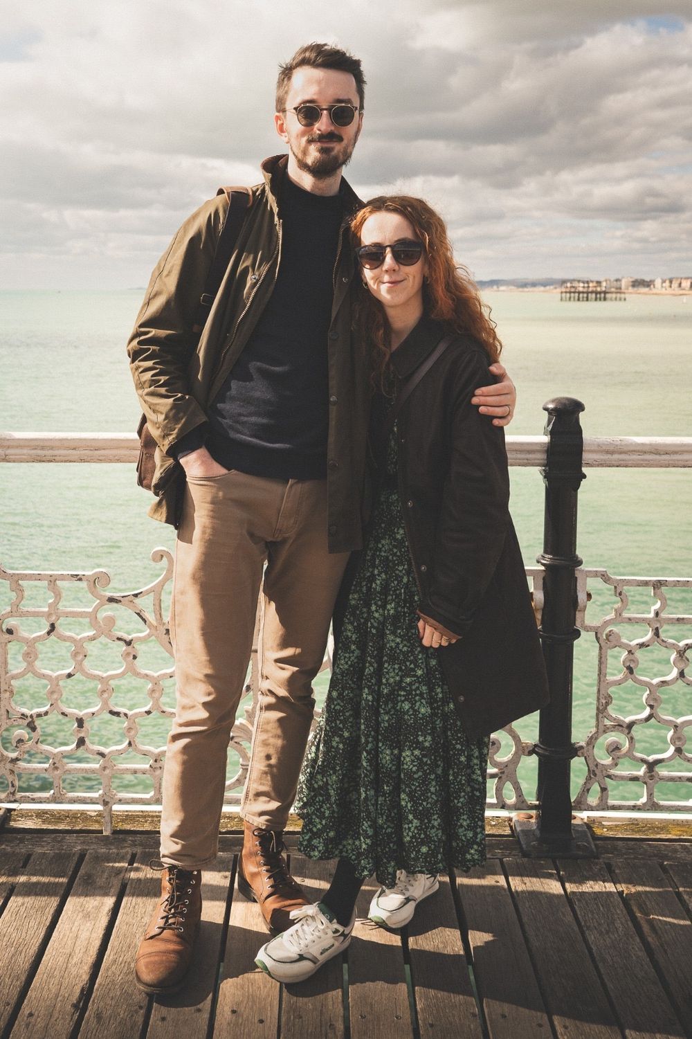 A couple stands on a wooden pier with ornate railings by the ocean under cloudy skies.