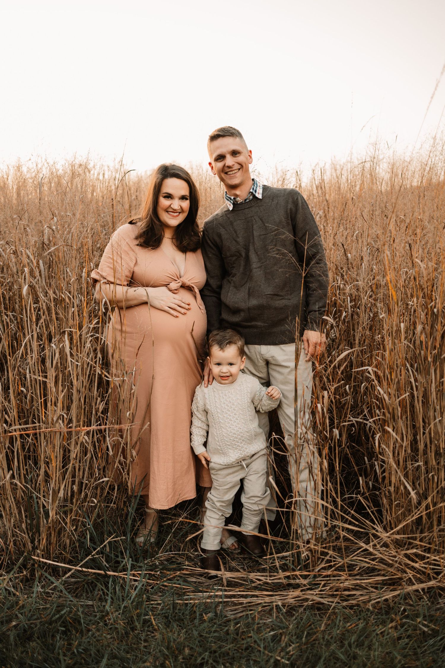 Pregnant woman in pink dress with husband and toddler standing in tall golden grass field at sunset.