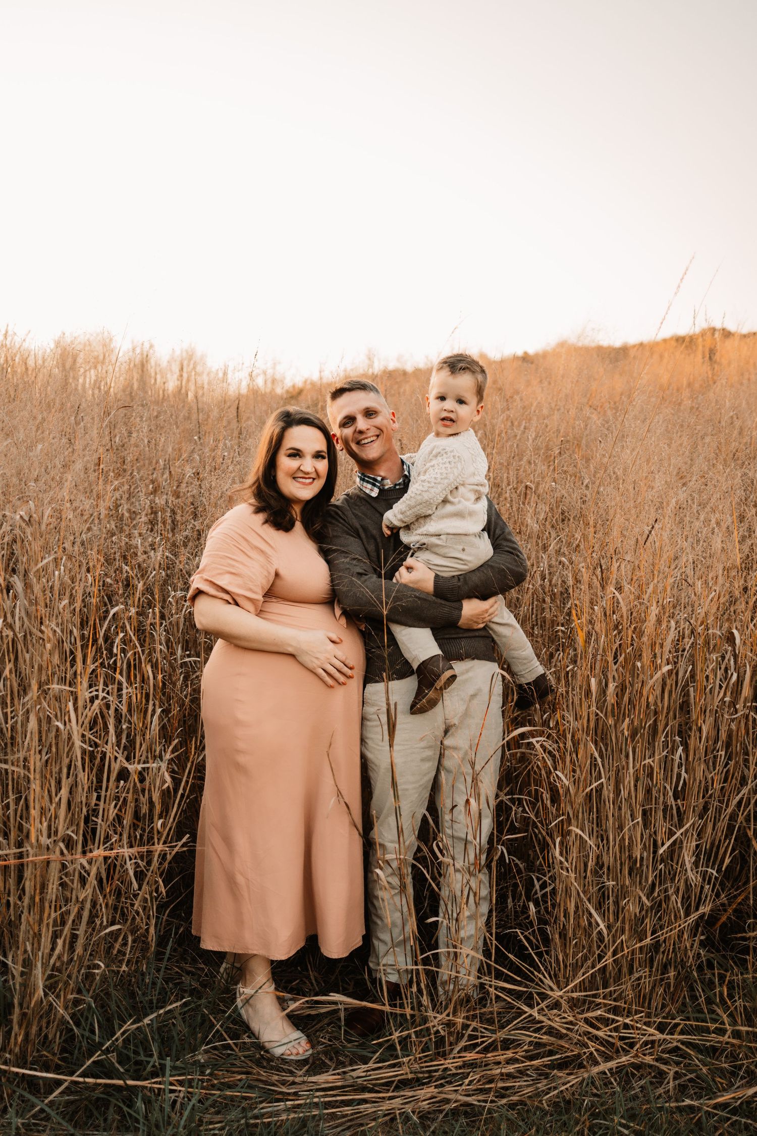 Smiling family with pregnant mother in pink dress, father holding toddler son in tall autumn grass field.