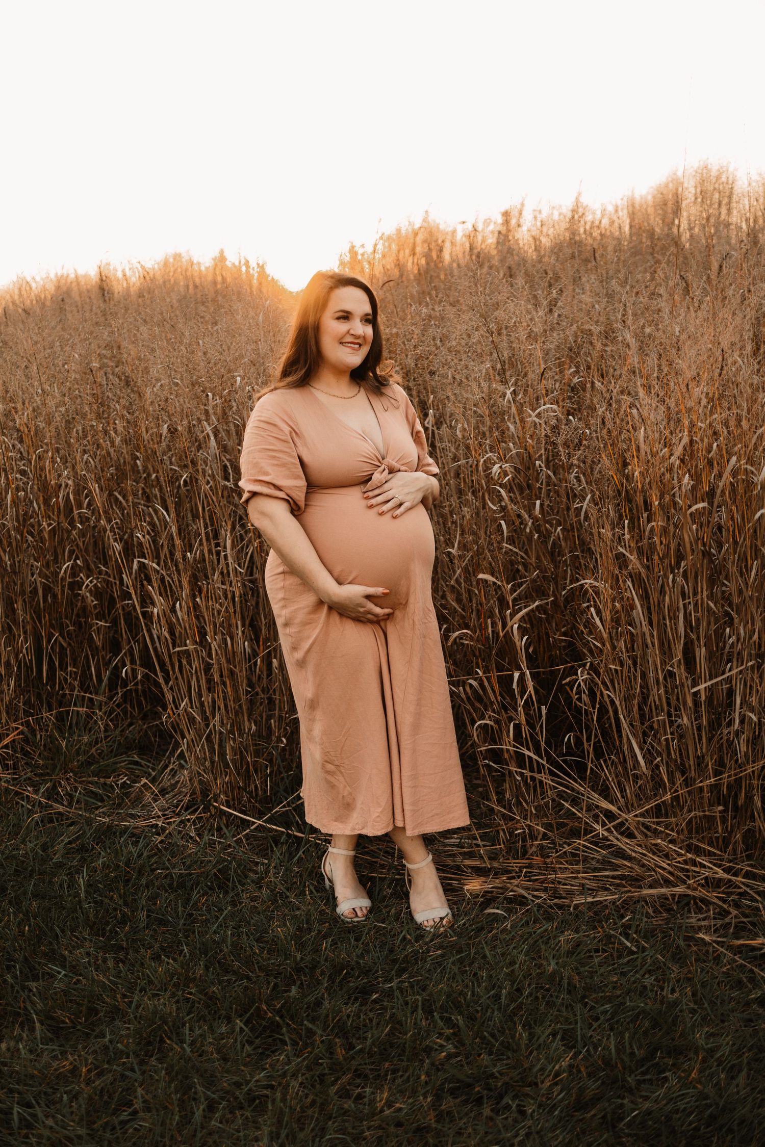 Glowing pregnant woman in blush pink dress cradling baby bump in tall golden grass at sunset.