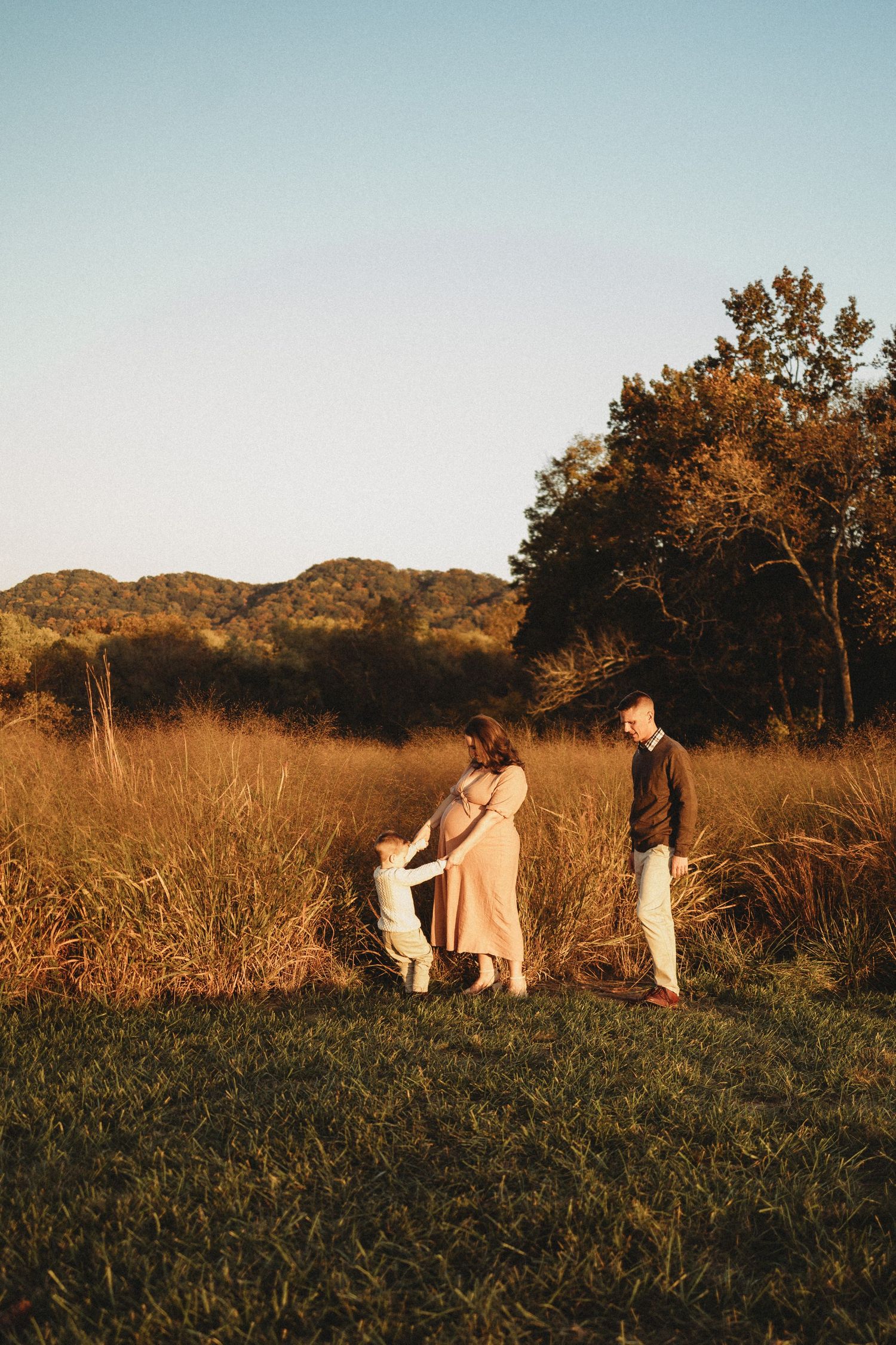 Family of three walking in sunlit field with tall golden grass and autumn trees in background.