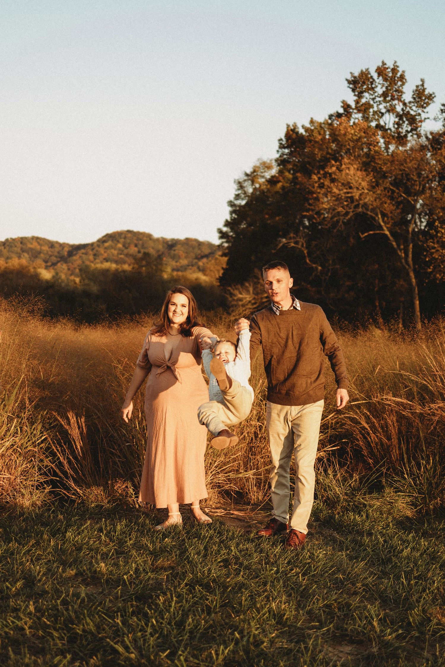 Happy family swinging toddler between pregnant mother and father in golden field at sunset.