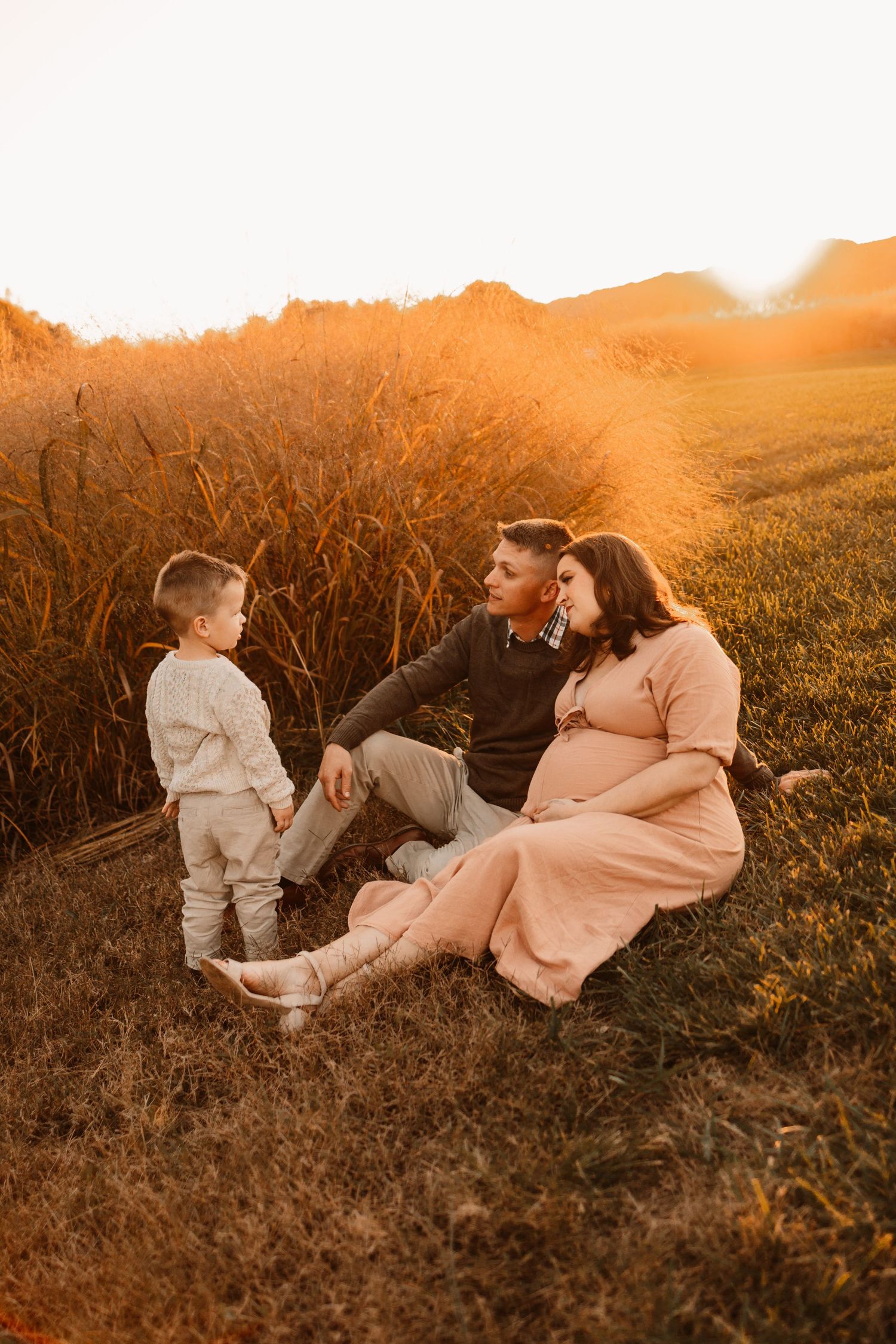 Family sitting together in warm golden sunset light beside tall grass field, toddler standing nearby.
