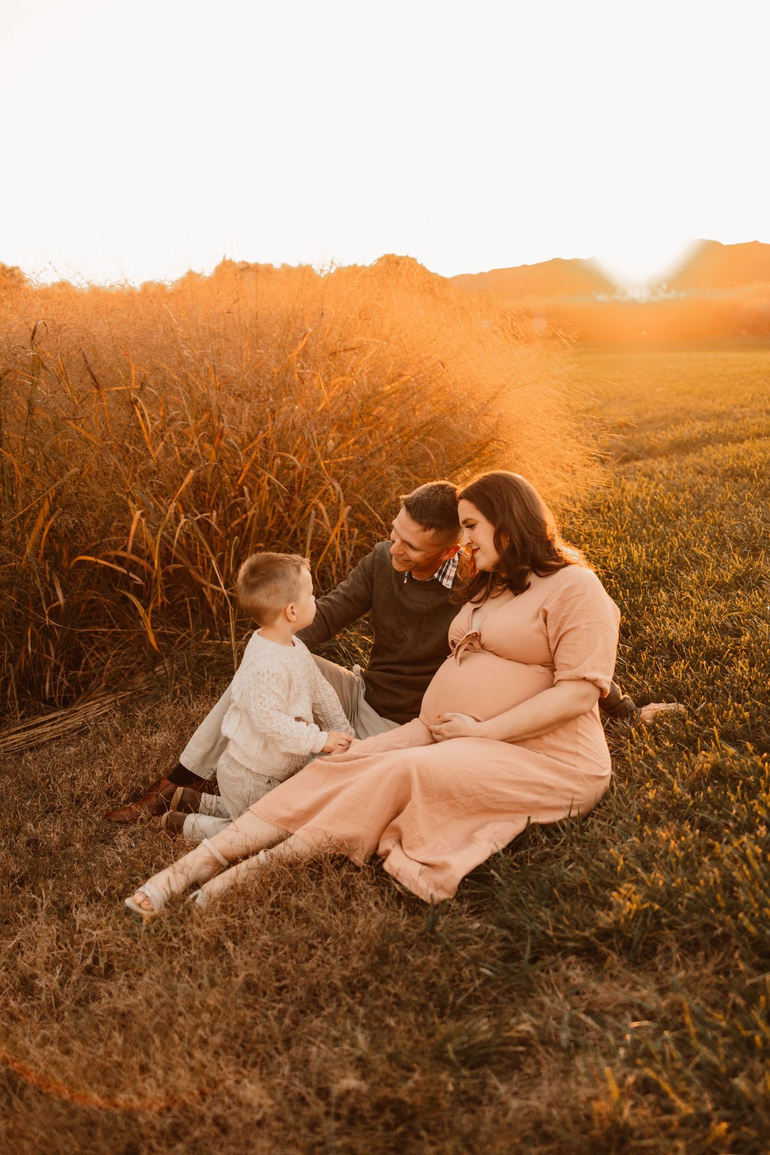 Couple sharing tender moment sitting in grass field at golden sunset with toddler beside them.