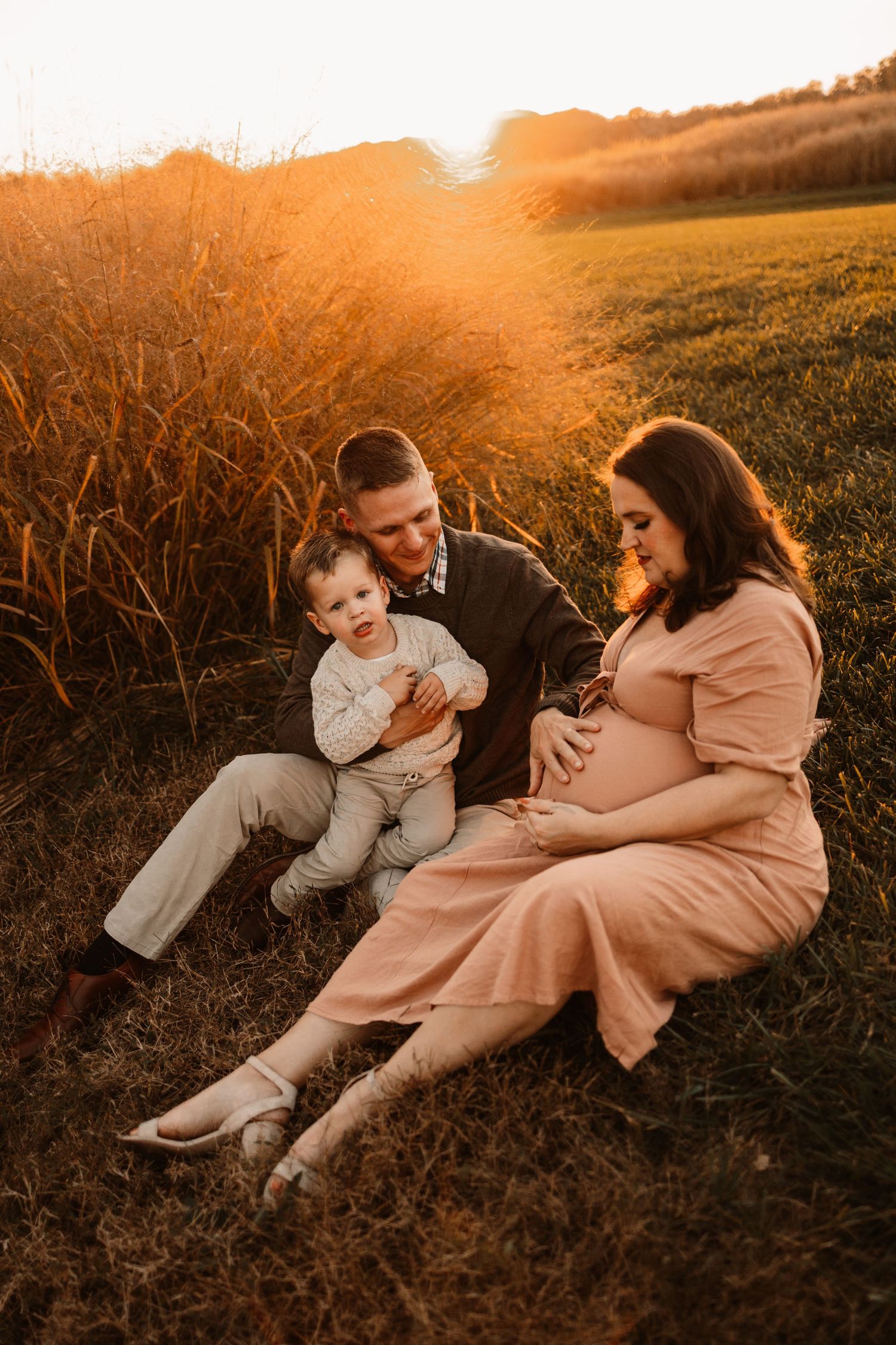 Family maternity photo at golden hour, couple sitting in grass with toddler beside tall wheat field.