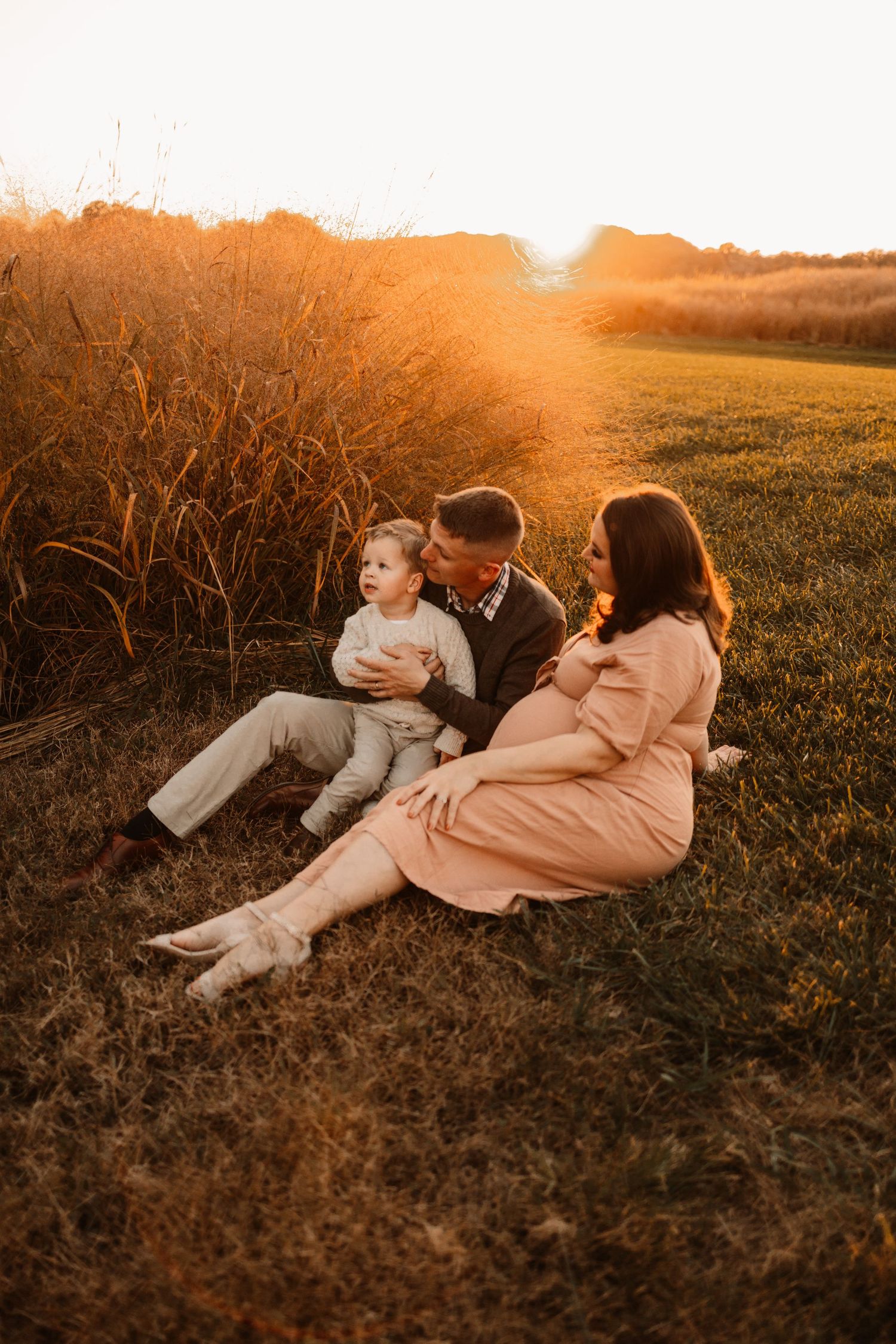 Couple sharing a kiss during golden hour maternity session while toddler sits between them outdoors.