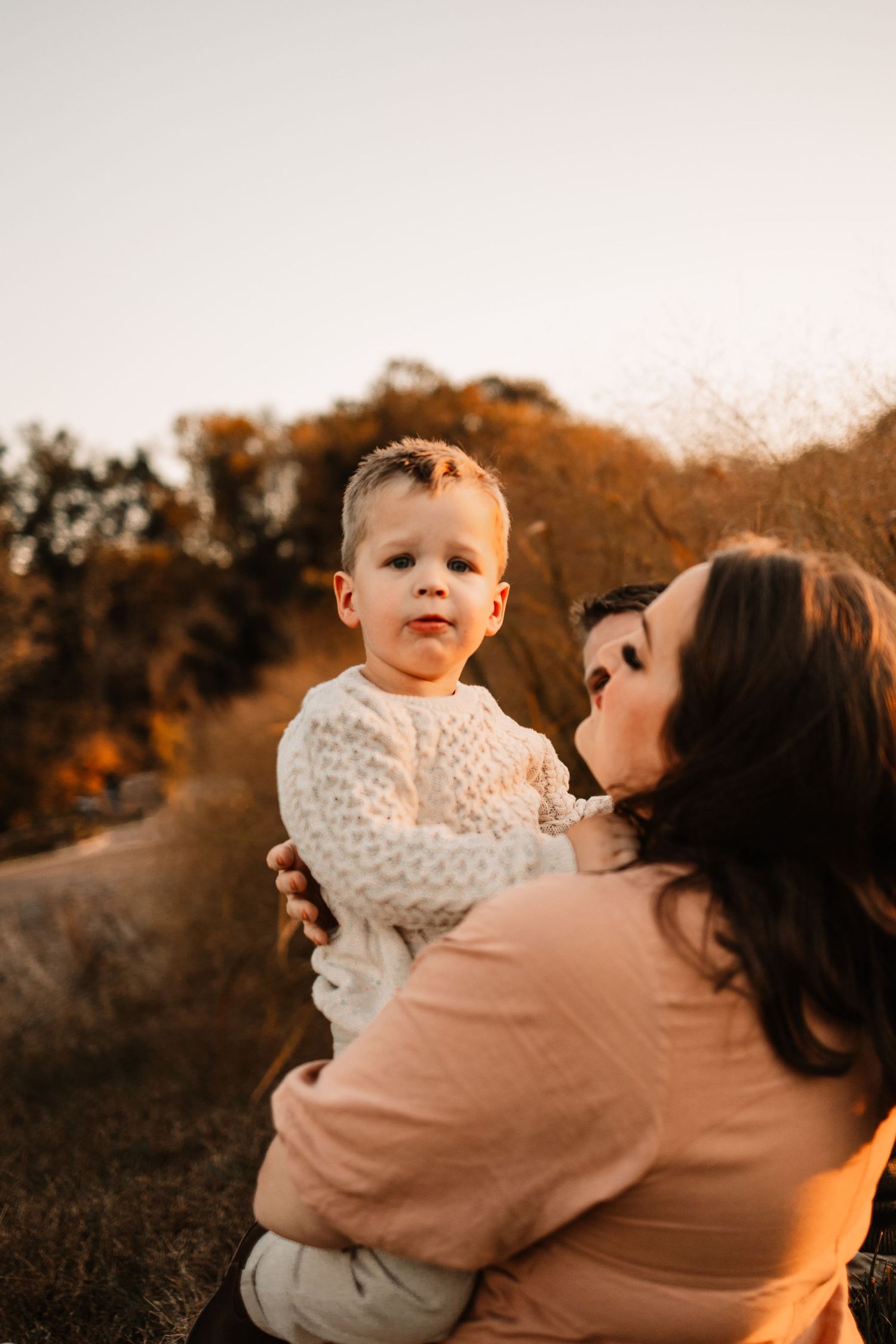 Mother holding young toddler boy in white sweater during golden hour outdoor maternity photoshoot.