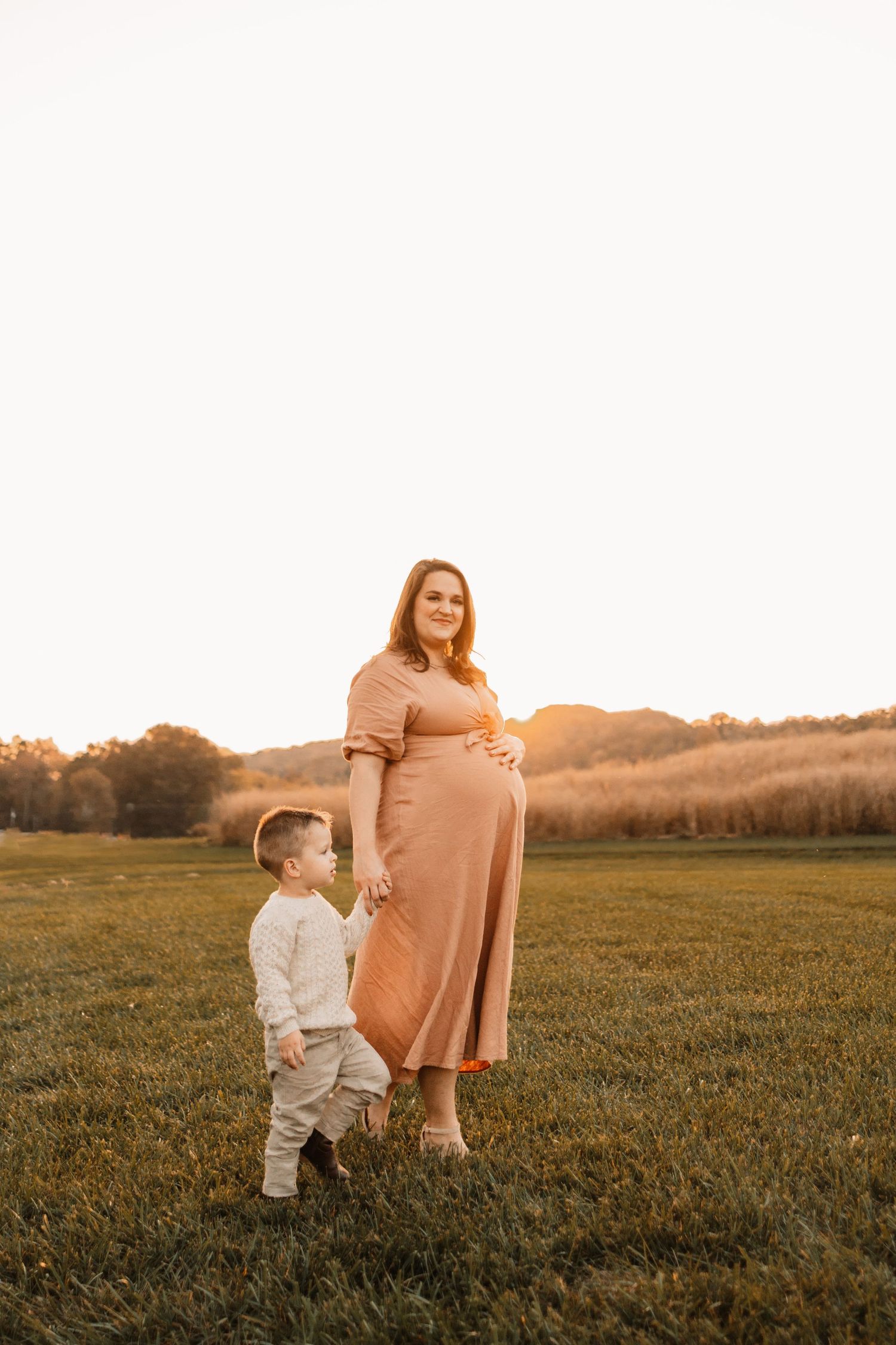 Pregnant woman in blush dress holds toddler son's hand in sunlit field during maternity session.