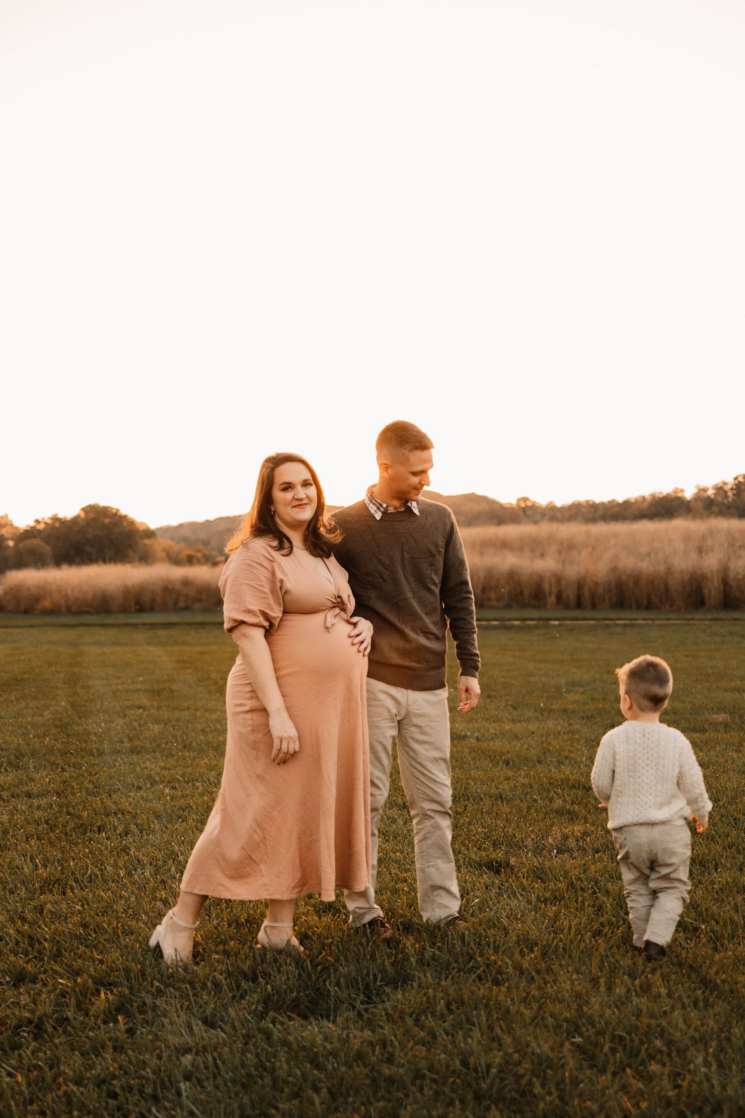 Pregnant couple walking in golden field at sunset with toddler son during fall maternity session.