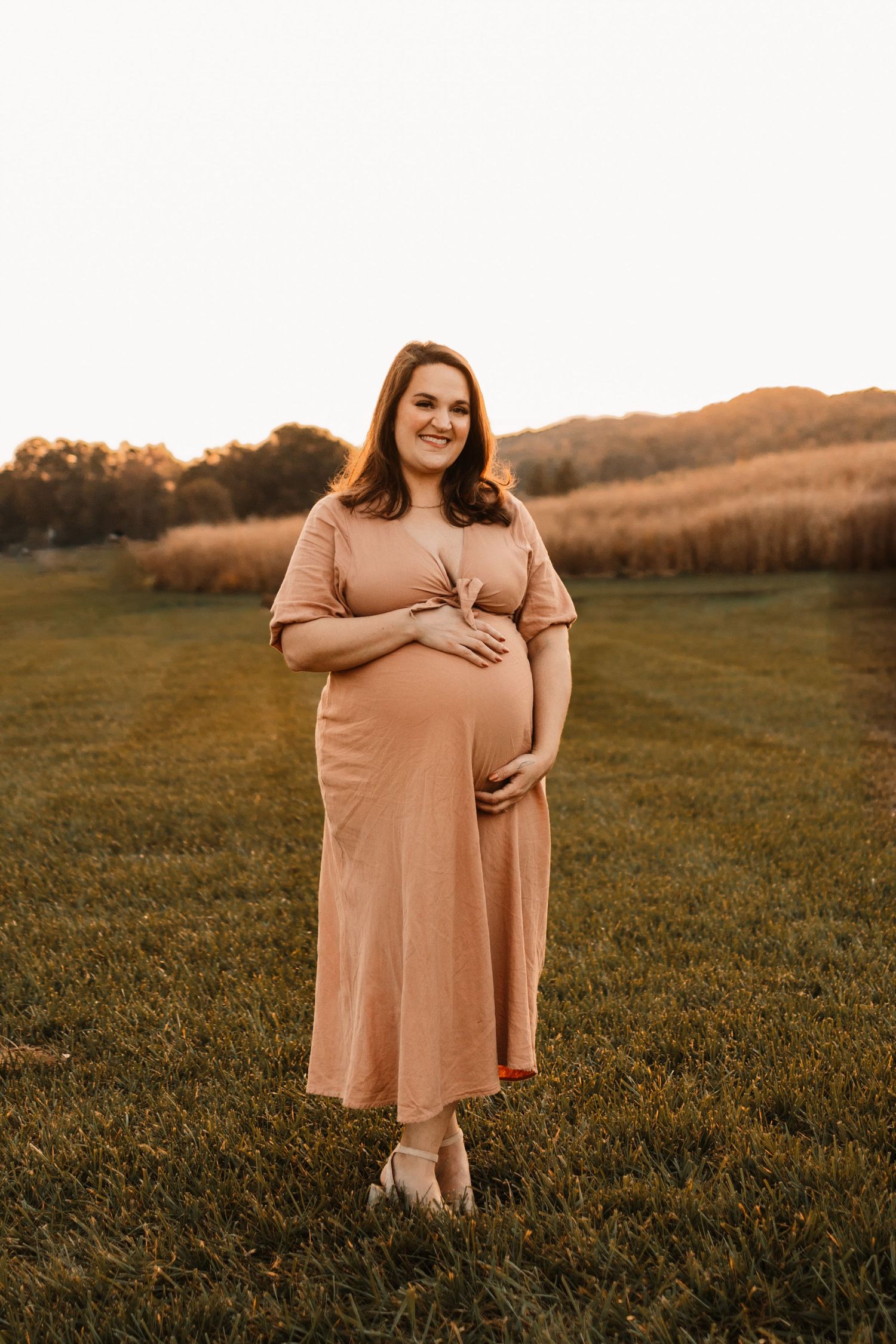 Smiling pregnant woman in blush midi dress cradles baby bump in sunlit field at golden hour.