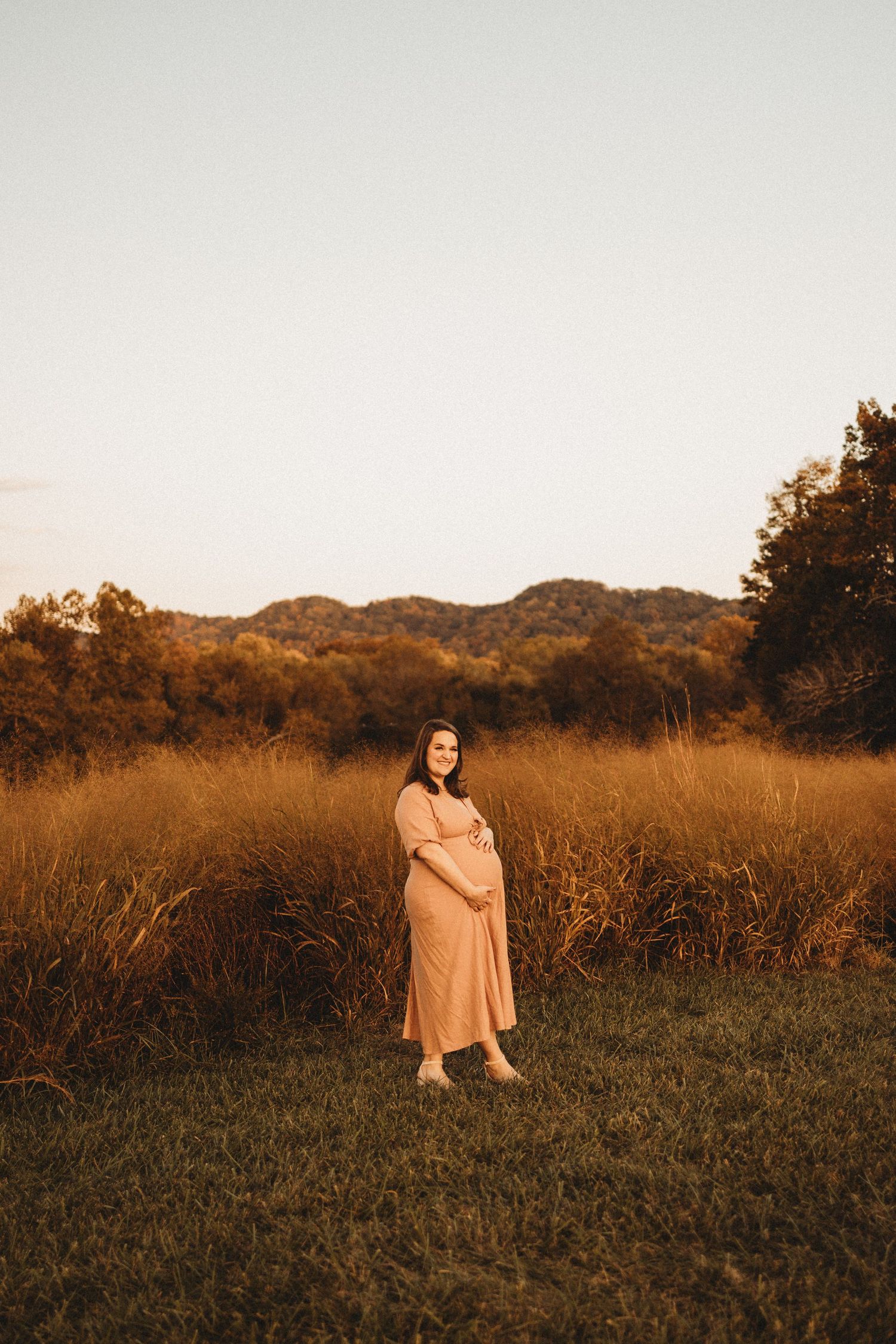 Pregnant woman in blush dress smiles while holding baby bump in golden hour field at sunset.