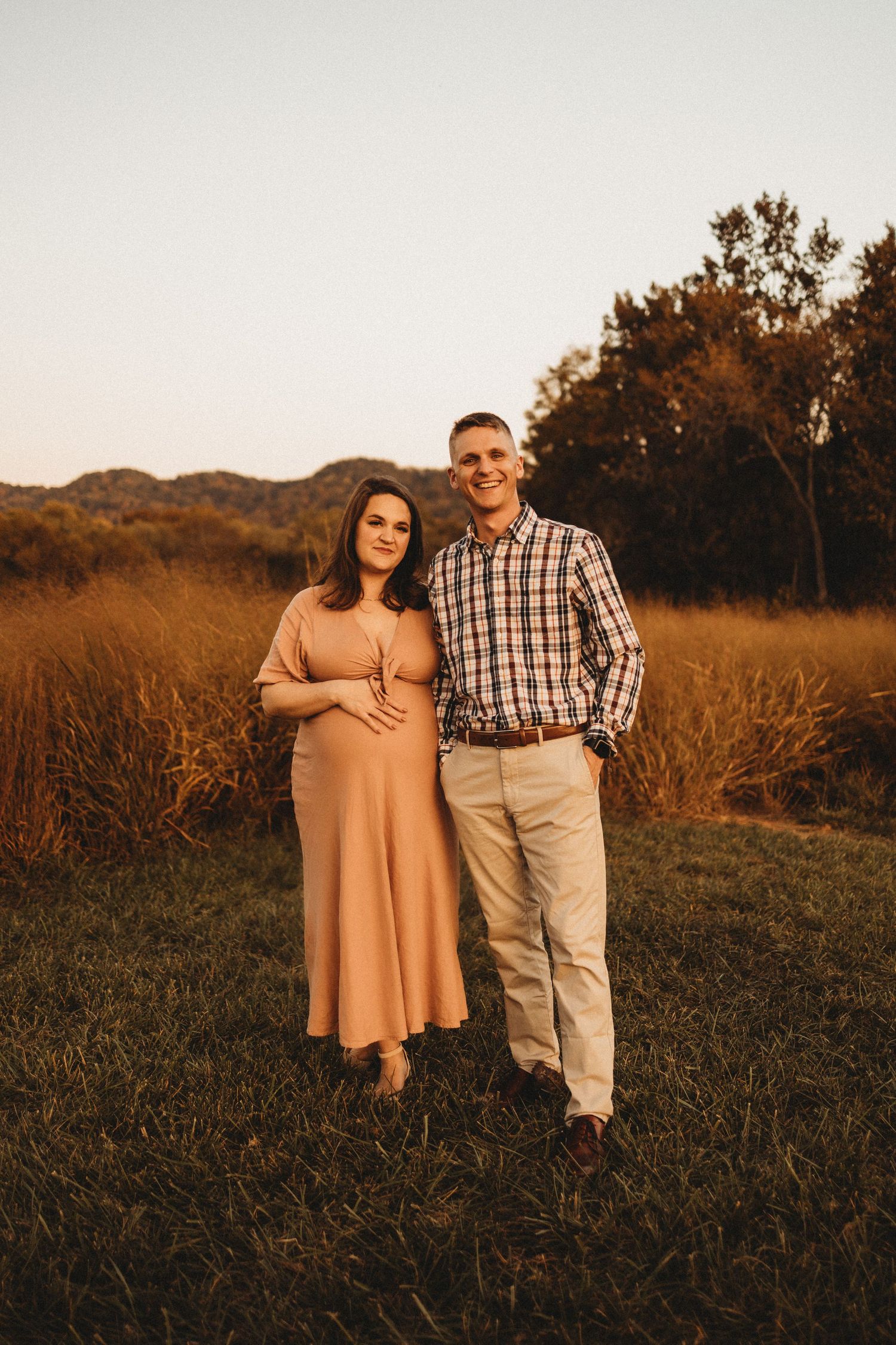 Smiling pregnant couple standing together in golden hour light during fall outdoor maternity session.