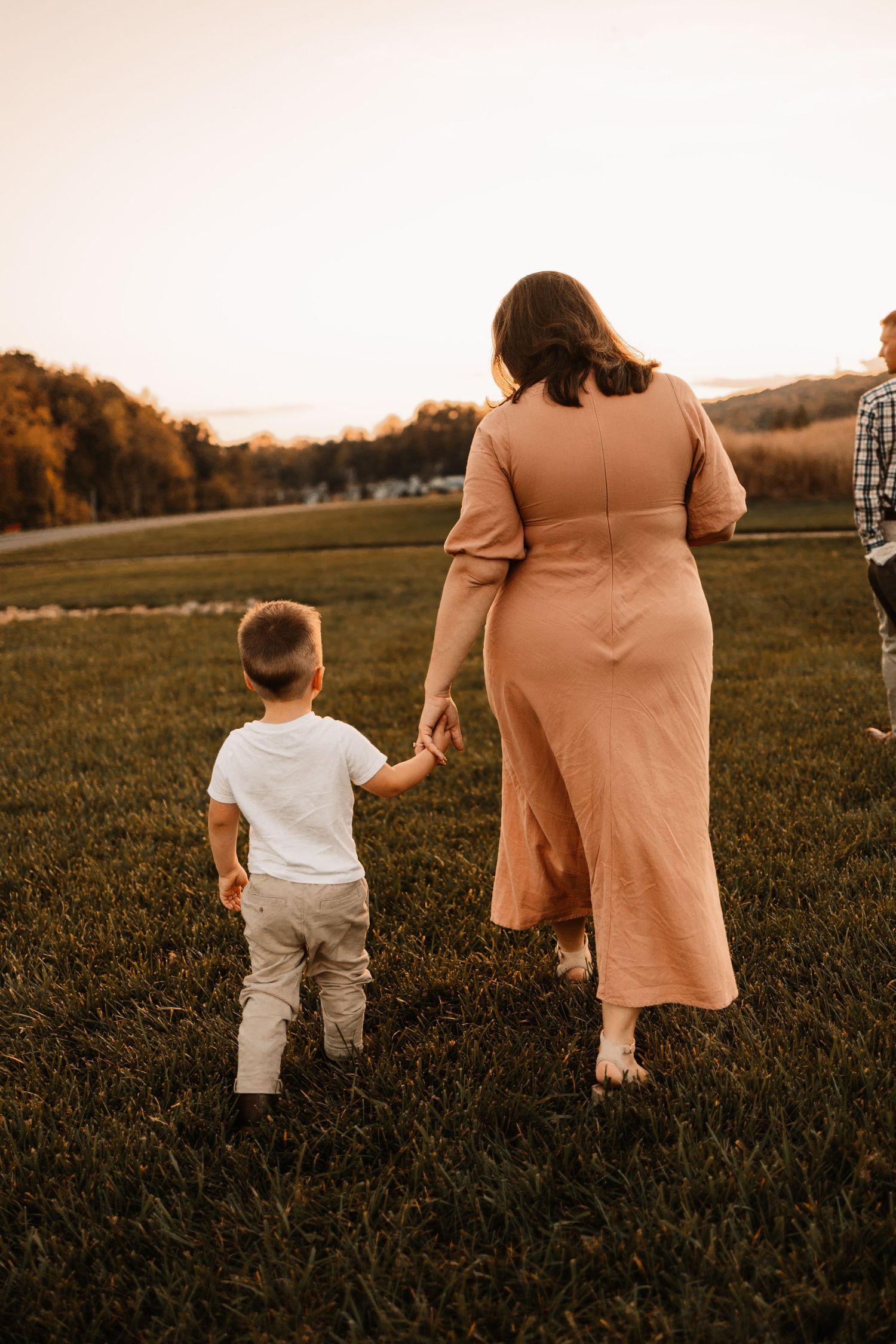 Pregnant mother in blush dress holding toddler son's hand walking through green field at golden hour sunset.