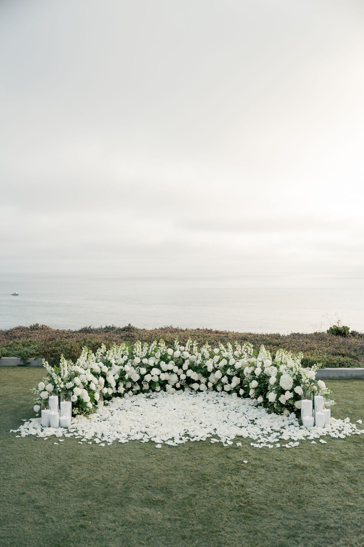 Outdoor wedding ceremony setup with elegant white floral arrangements and decor along a curved path by the ocean.