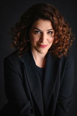 Professional headshots in black blazer with curly hair against dark backdrop.