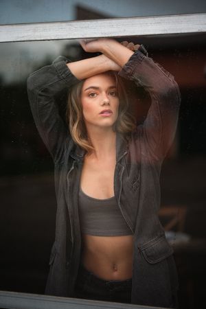 Moody portrait of a woman in crop top and jacket posing by a window.