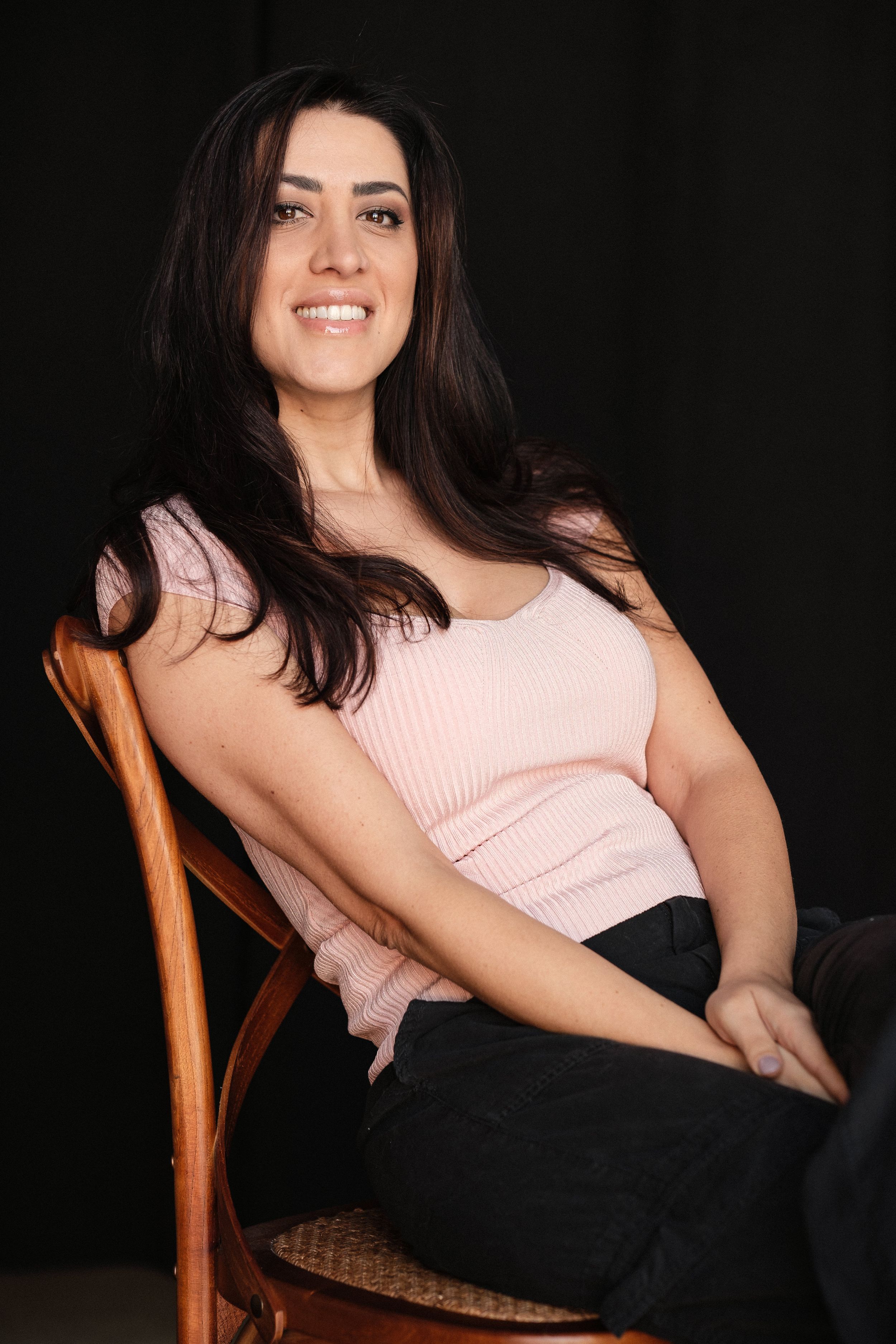 Studio portraits featuring pink top and relaxed poses in wooden chair against dark background.