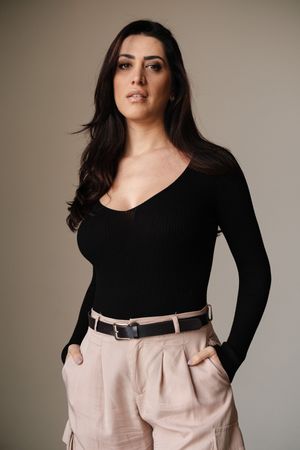 Fashion poses in black top and beige pants with belt against neutral background.