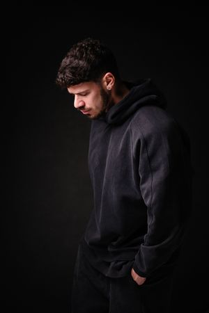 Moody portrait of person in black hoodie looking down against dark background.